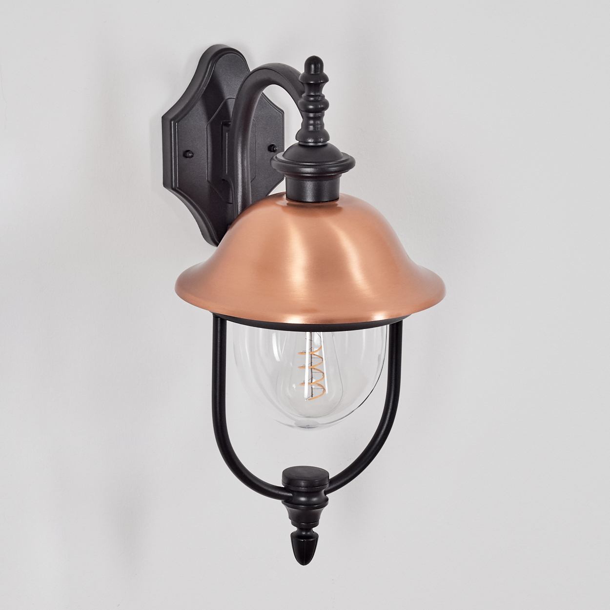 Eine Außenwandleuchte. Sie hat einen kupferfarbenen Schirm und einen schwarzen Rahmen. Die Glühbirne ist sichtbar. Die Lampe ist an einer weißen Wand.