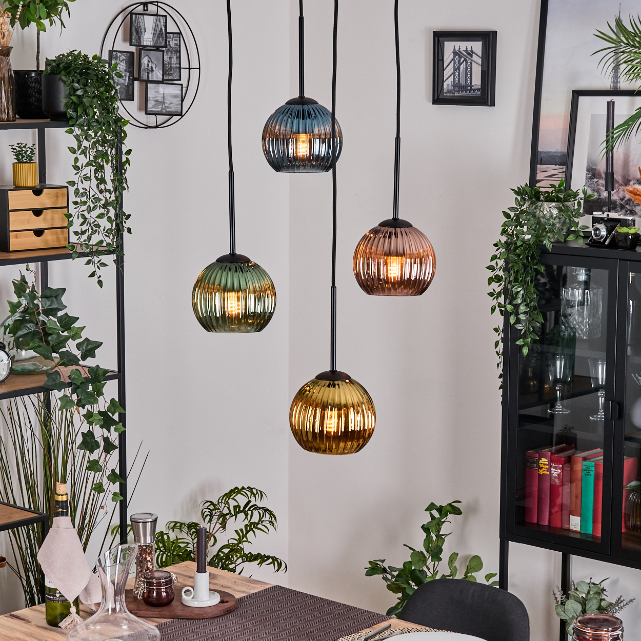 Vier bunte Pendelleuchten über einem Holztisch in einem Esszimmer. Pflanzen und Regale sind im Hintergrund.