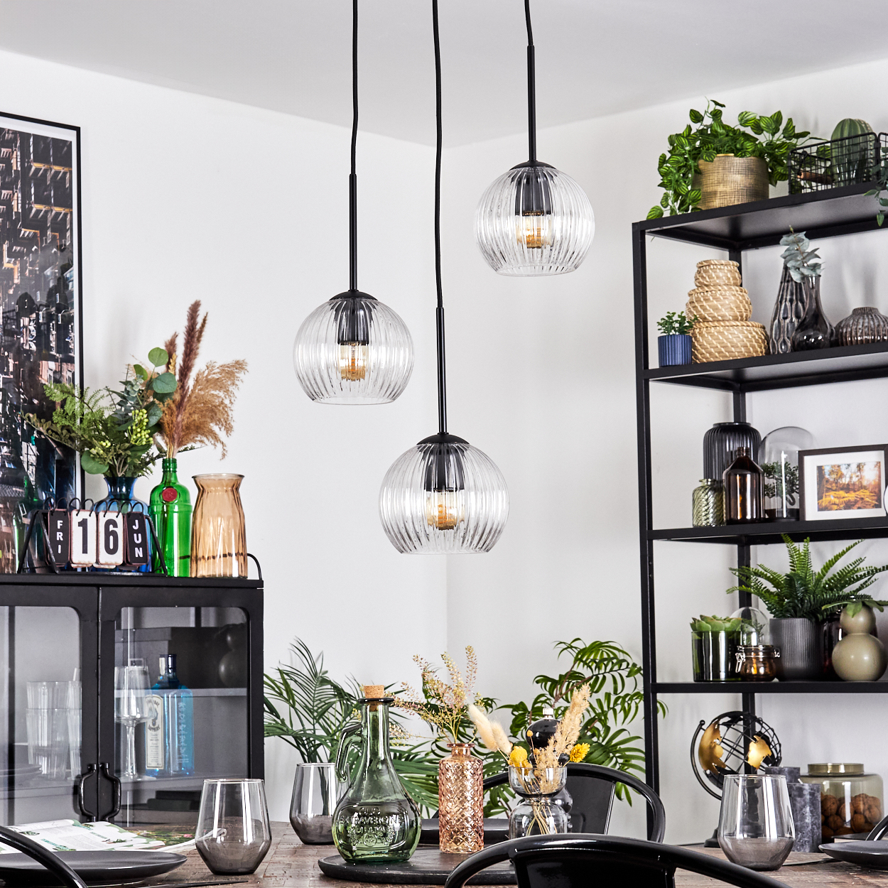 Esszimmer mit drei Hängelampen. Tisch mit Gläsern und Dekoration. Regale mit Pflanzen und Vasen im Hintergrund.