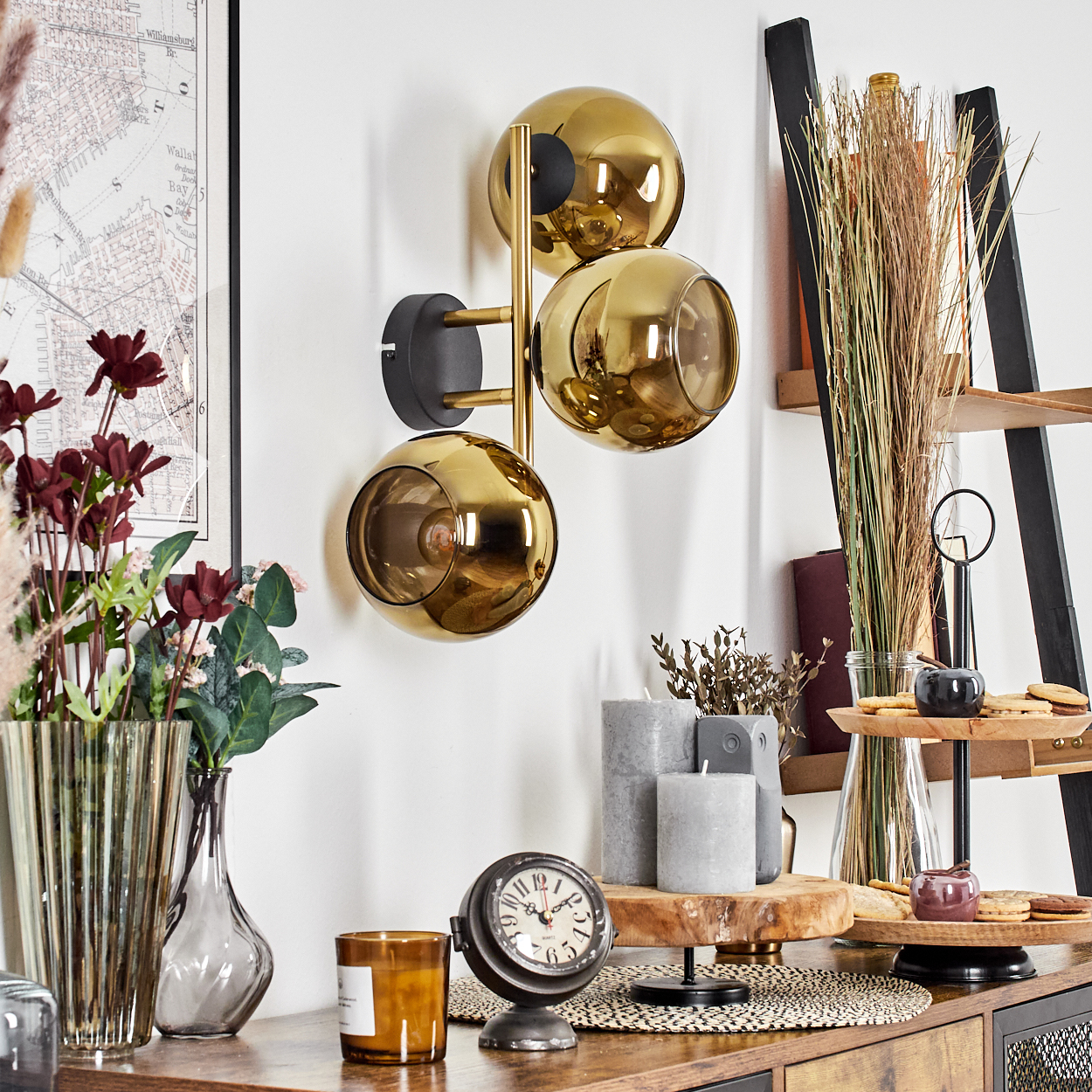 Eine goldene Kugellampe hängt an einer weißen Wand. Auf einem Holztisch stehen Blumen, eine Uhr, Kerzen und ein Tablett mit Keksen.