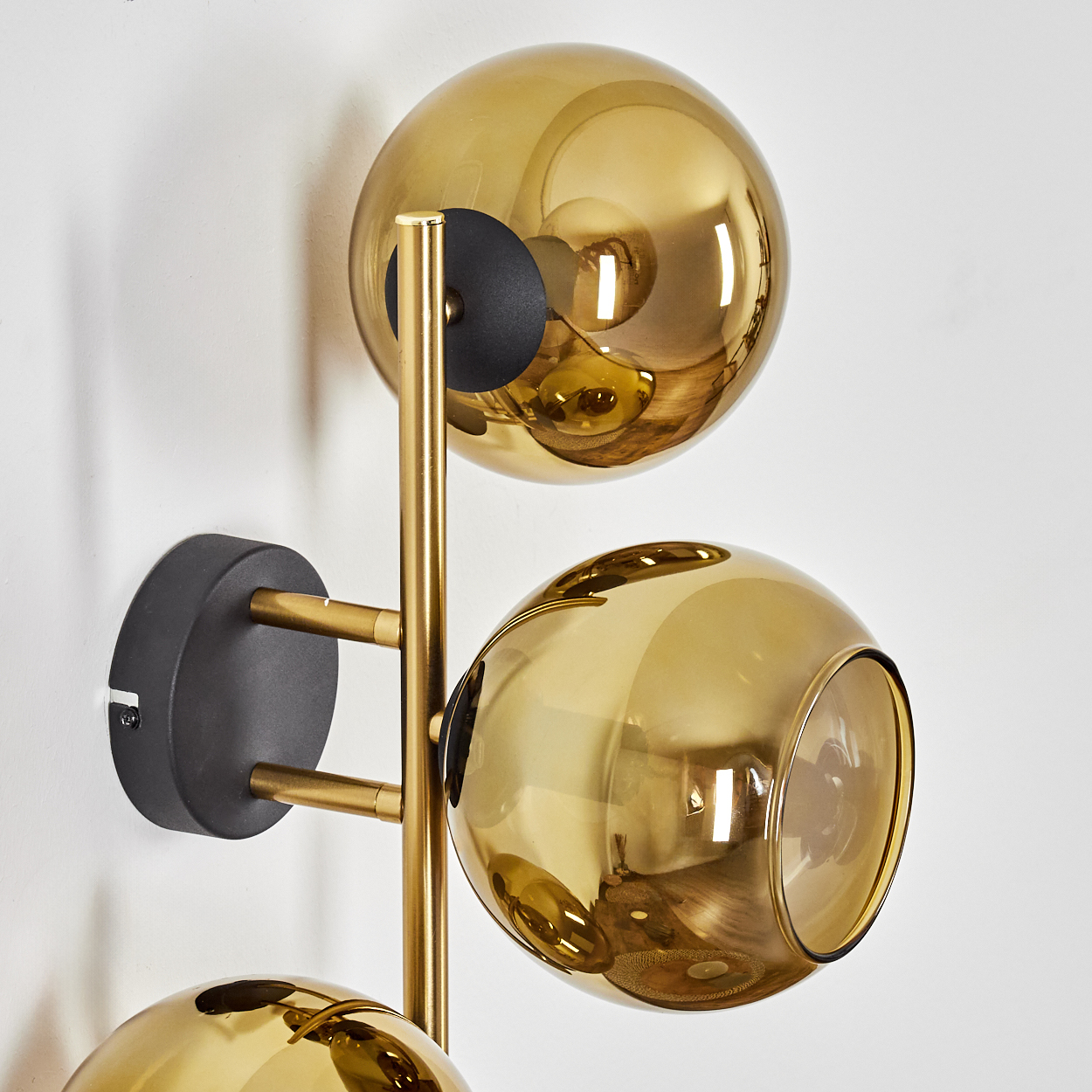 Eine moderne Wandleuchte mit drei goldenen Glaskugeln und goldenem Metallrahmen an einer schwarzen Basis an einer weißen Wand.