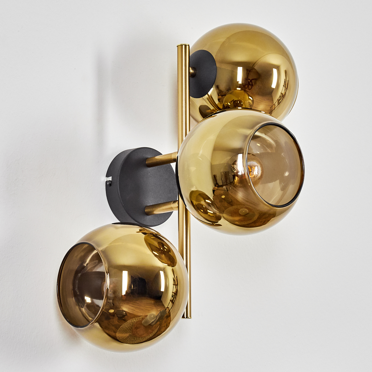 Eine moderne Wandleuchte mit drei goldenen kugelförmigen Schirmen, die an einer schwarzen Basis befestigt sind, auf weißer Wand.