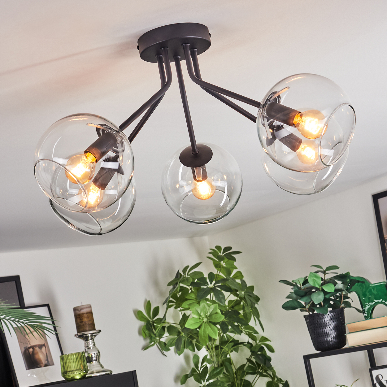 Eine moderne Deckenleuchte mit drei Glaskugeln. Das Licht ist an und beleuchtet den Raum. Pflanzen und Dekorationen sind im Hintergrund.