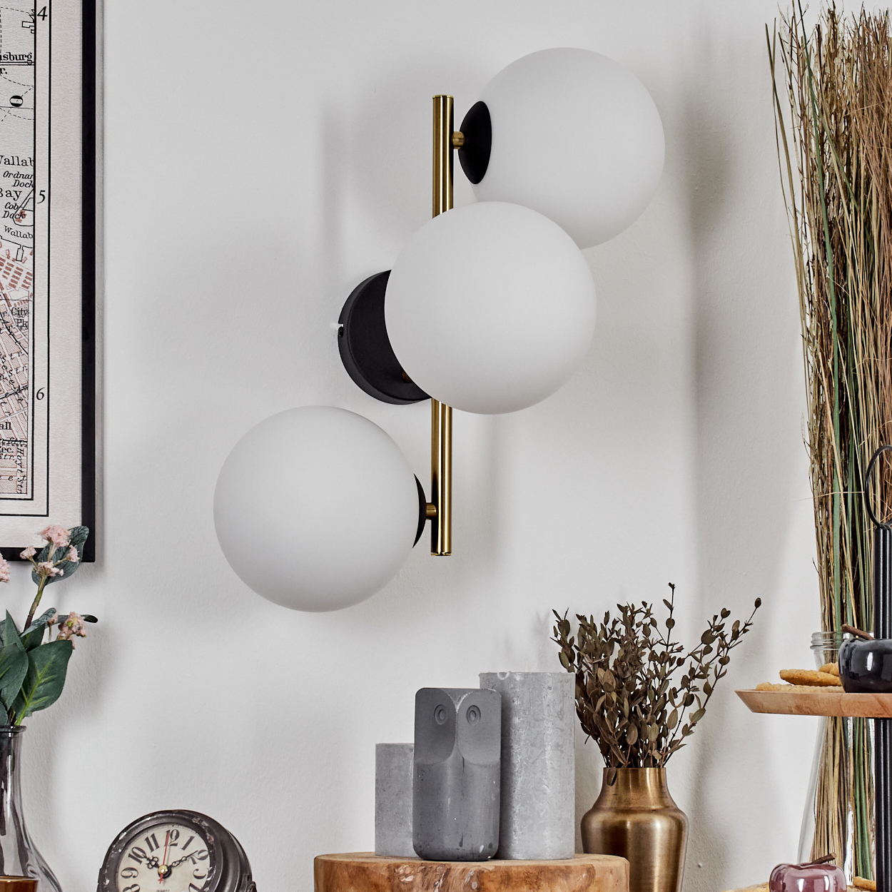 Eine Wandlampe mit drei weißen Kugeln ist an einer weißen Wand befestigt. Darunter zeigt ein Tisch Dekorationsartikel: eine Eule, eine Uhr und eine Vase.
