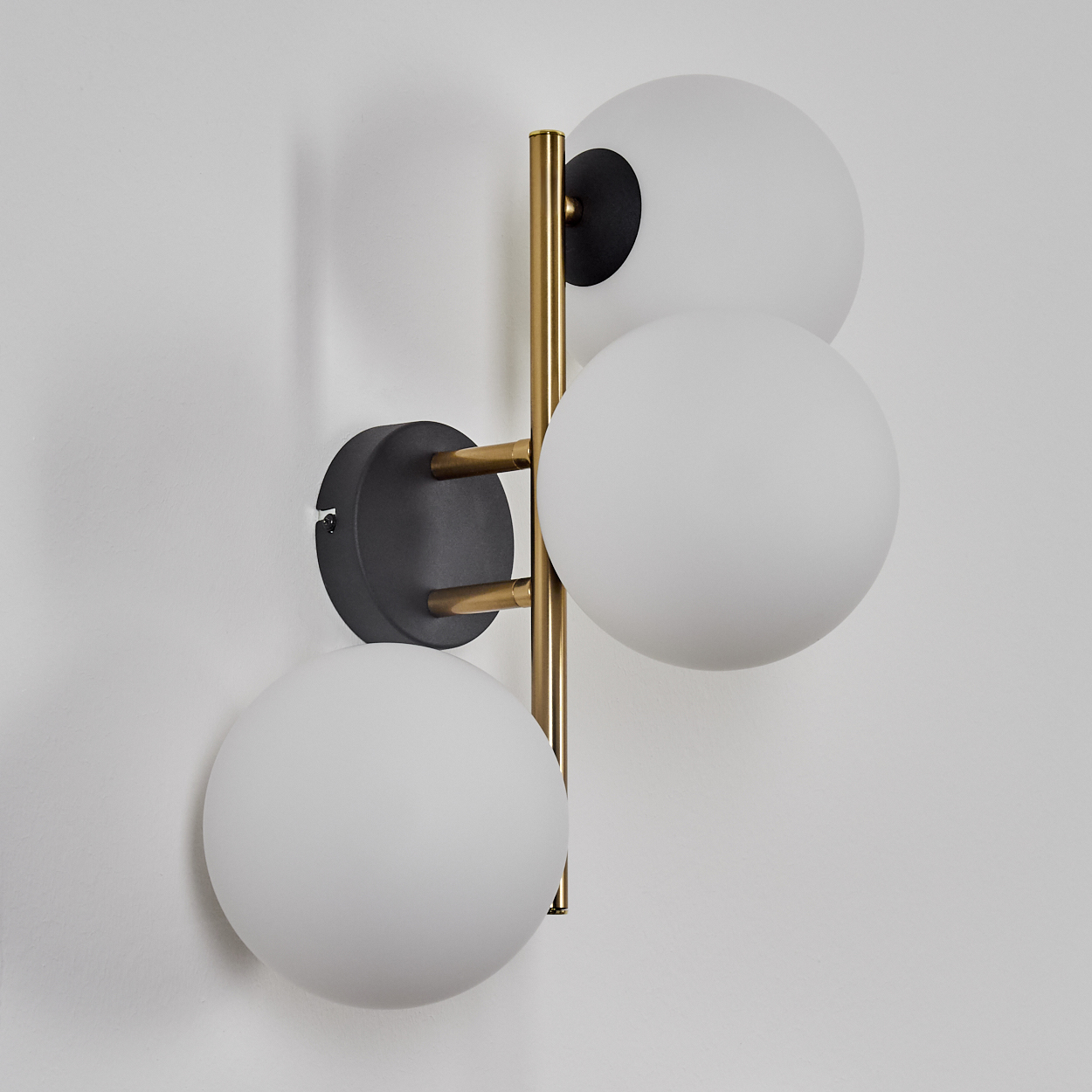Eine moderne Wandlampe mit vier weißen Kugeln. Sie hat einen schwarzen Sockel und ein goldfarbenes Metallgestell, montiert an einer weißen Wand.