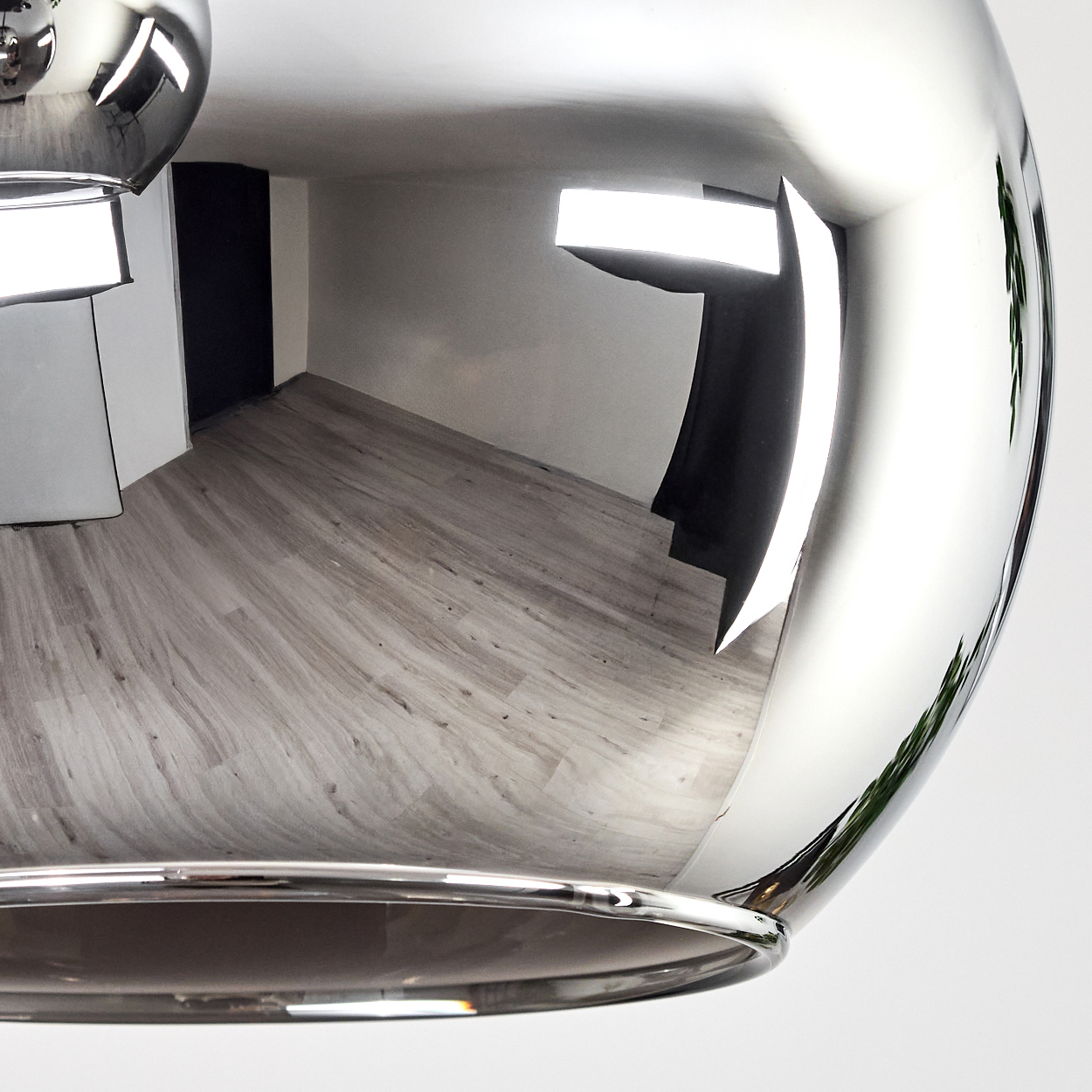 Eine verchromte, reflektierende Kugel, die einen Raum mit grauem Holzboden, weißen Wänden und einer schwarzen Ecke reflektiert. Helle Lichter werden ebenfalls reflektiert.