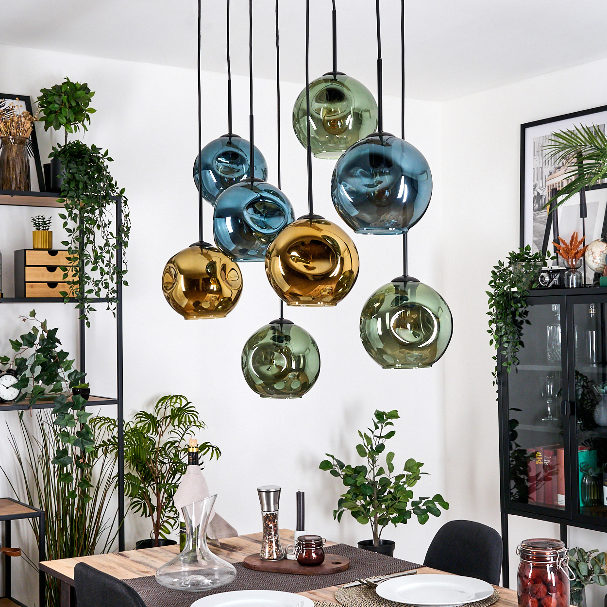 Esszimmer mit Holztisch, hängenden Kugelleuchten und Pflanzen. Goldene, blaue und grüne Glasleuchten. Regale und ein Schrank.