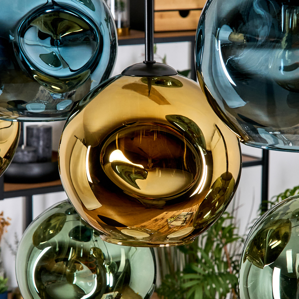 Nahaufnahme von gläsernen Pendelleuchten in Gold-, Blau- und Grüntönen. Die Glühbirnen reflektieren die Umgebung. Grüne Pflanzen im Hintergrund.