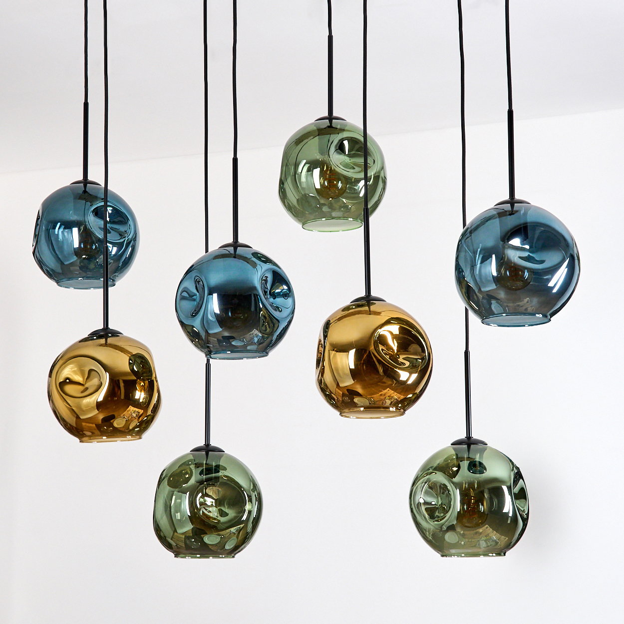 Acht kugelförmige Pendelleuchten. Farben sind Blau, Grün und Gold. Schwarze Kabel hängen von der Decke vor weißem Hintergrund.