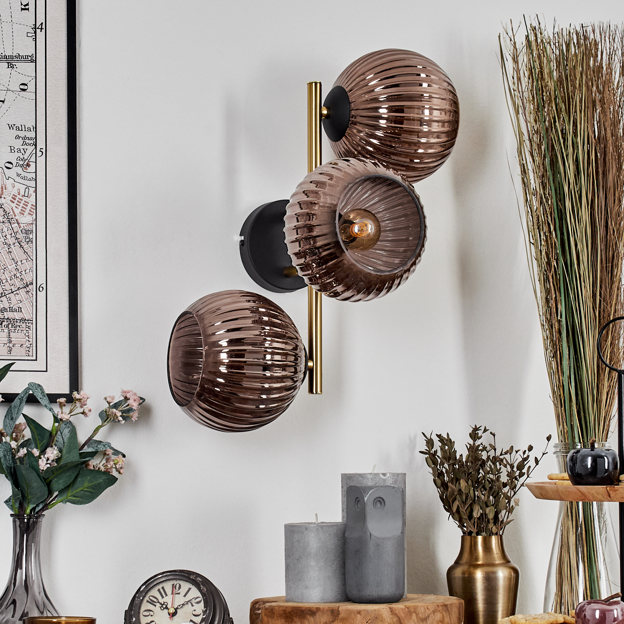 Eine Wandlampe mit drei braunen Glaskugeln. Darunter Kerzen, eine Uhr und eine Vase mit Blumen. Im Hintergrund Karte und Dekoration.