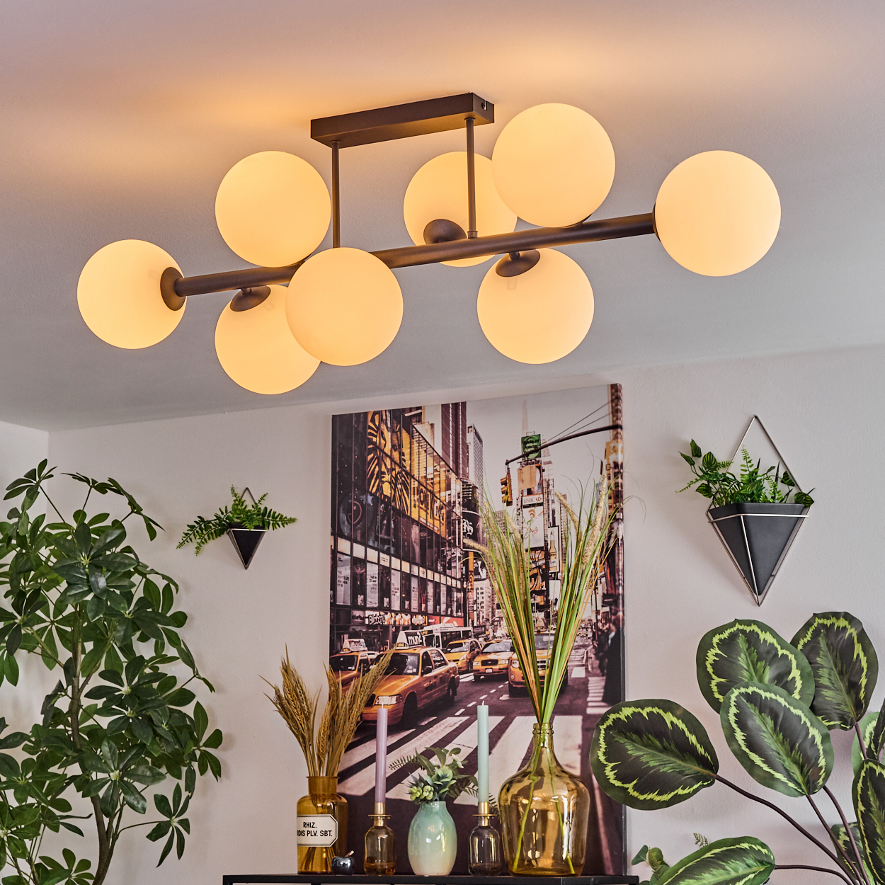 Ein modernes Wohnzimmer mit einem großen kugelförmigen Kronleuchter, Kunstwerken einer Stadtszene und verschiedenen Pflanzen. Ein schwarzes Regal mit Vasen.