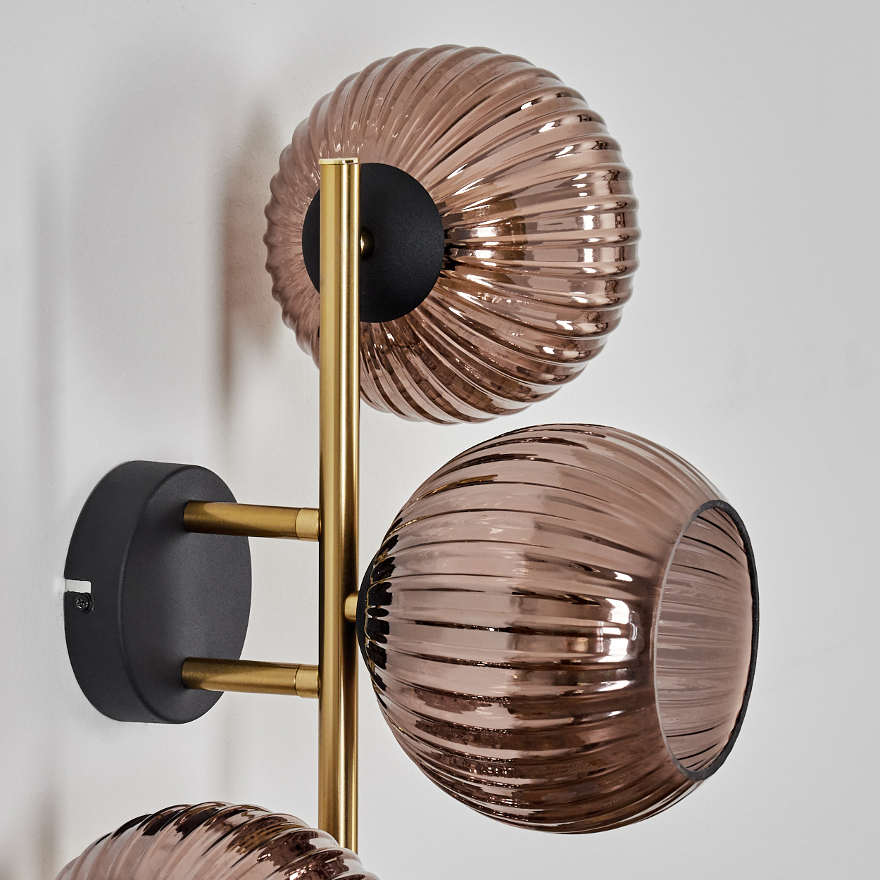 Eine moderne Wandlampe mit drei braunen Glaskugeln, goldenen Metallarmen und einem schwarzen Sockel vor weißer Wand.
