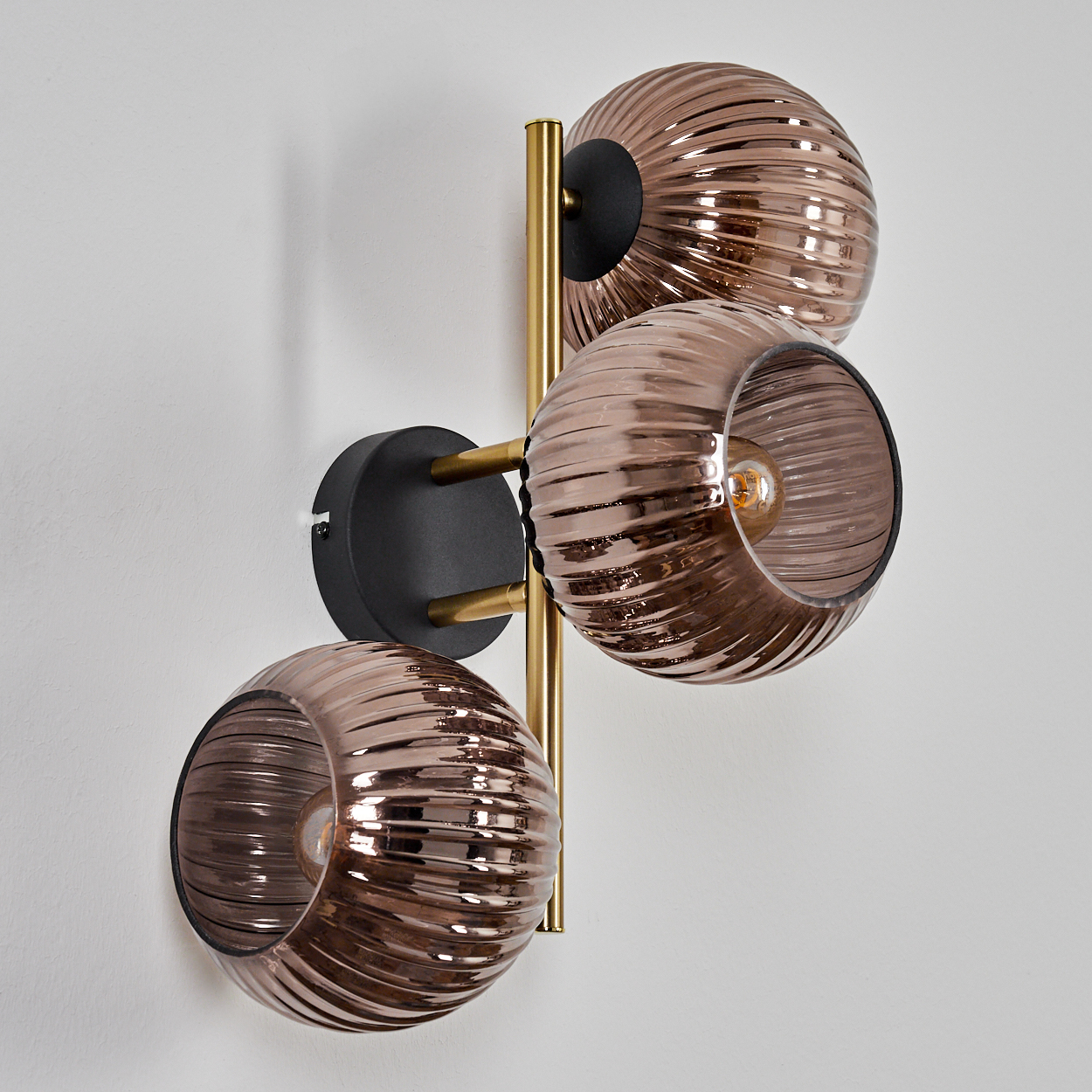 Eine moderne Wandlampe mit drei braunen Glaskugeln, goldenen Metallarmen und einem schwarzen Sockel, an einer weißen Wand montiert.