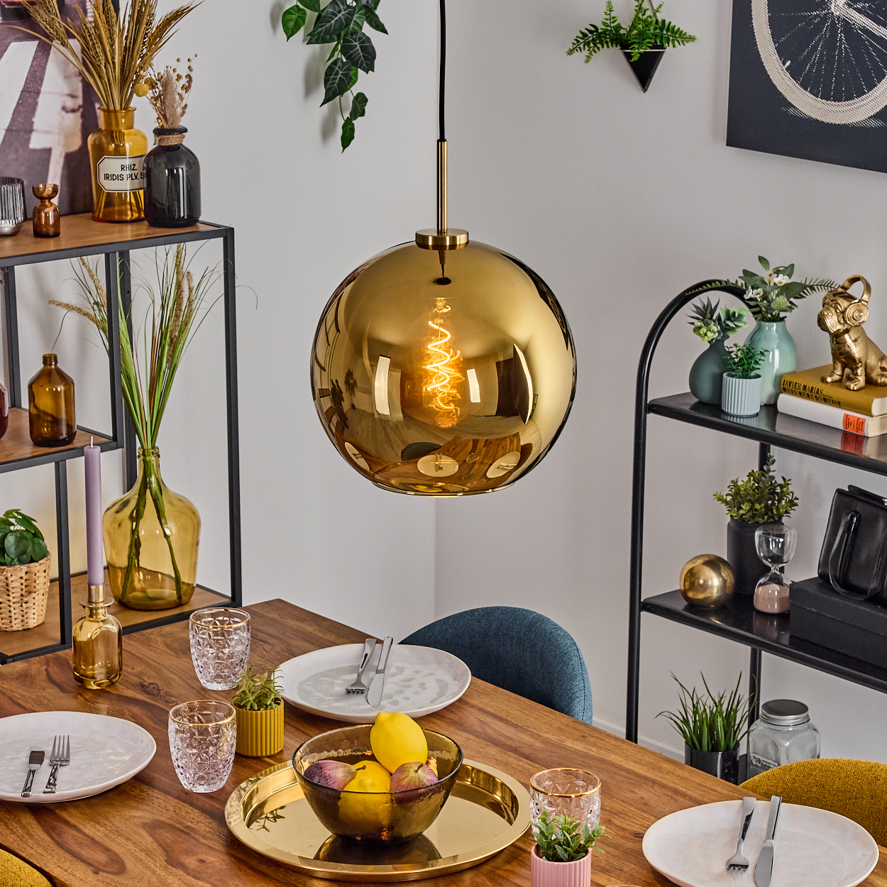 Ein Esstisch mit goldener Pendelleuchte, gedeckt mit Tellern, Gläsern und einer Obstschale. Regale und Pflanzen sind im Hintergrund.