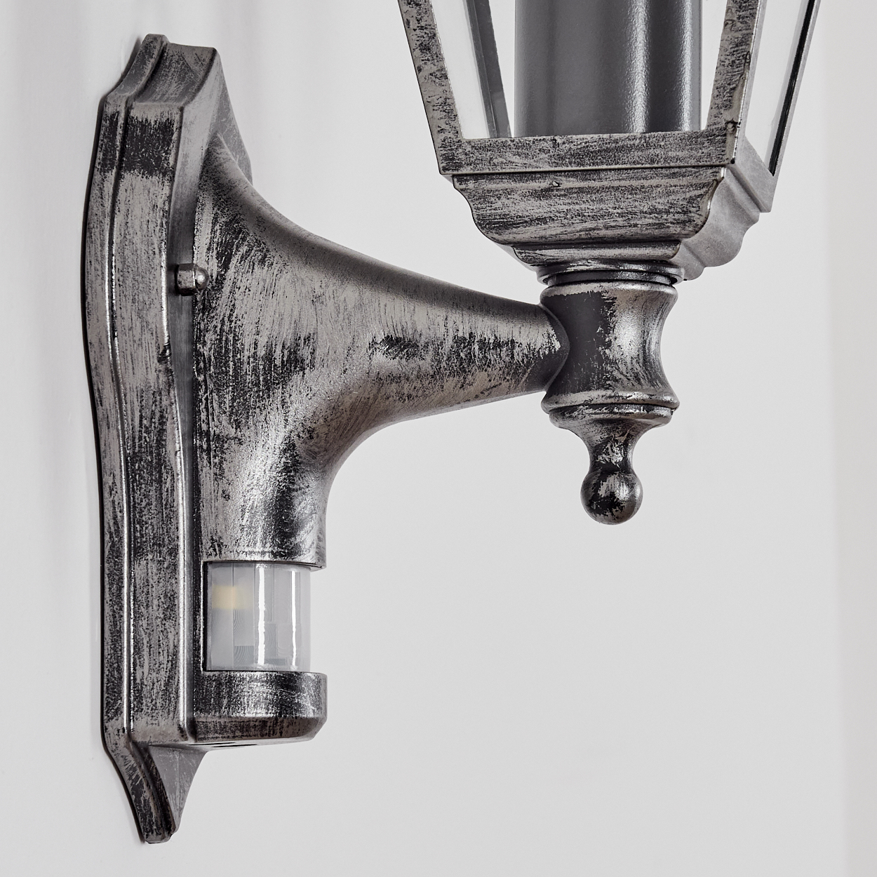 Eine silberne Außenlampe mit Bewegungssensor ist an einer weißen Wand befestigt. Die Lampe hat ein klassisches Design.