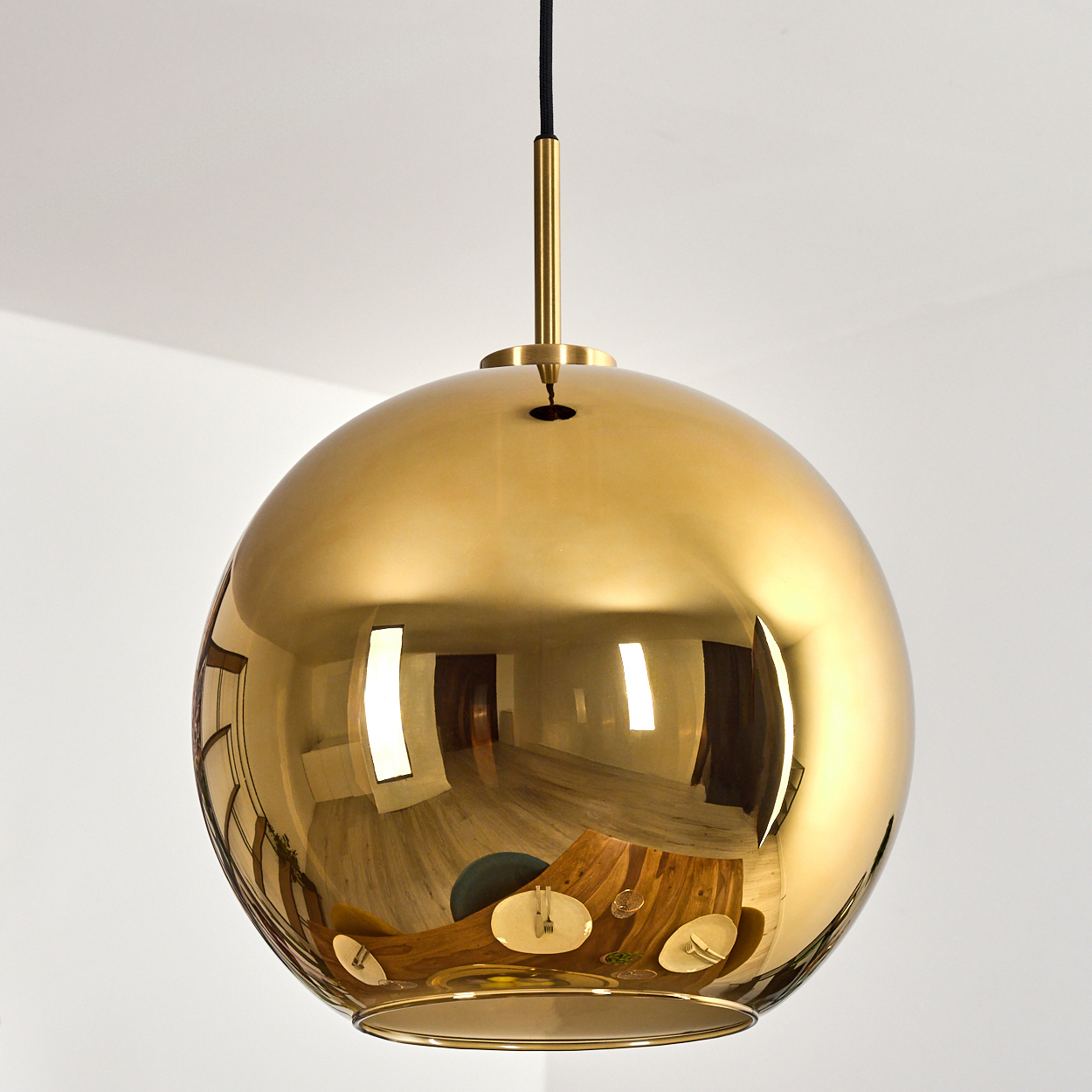Eine goldene, kugelförmige Hängelampe hängt von der Decke. Sie reflektiert einen Raum mit einem Holztisch, Stühlen und weißen Wänden.