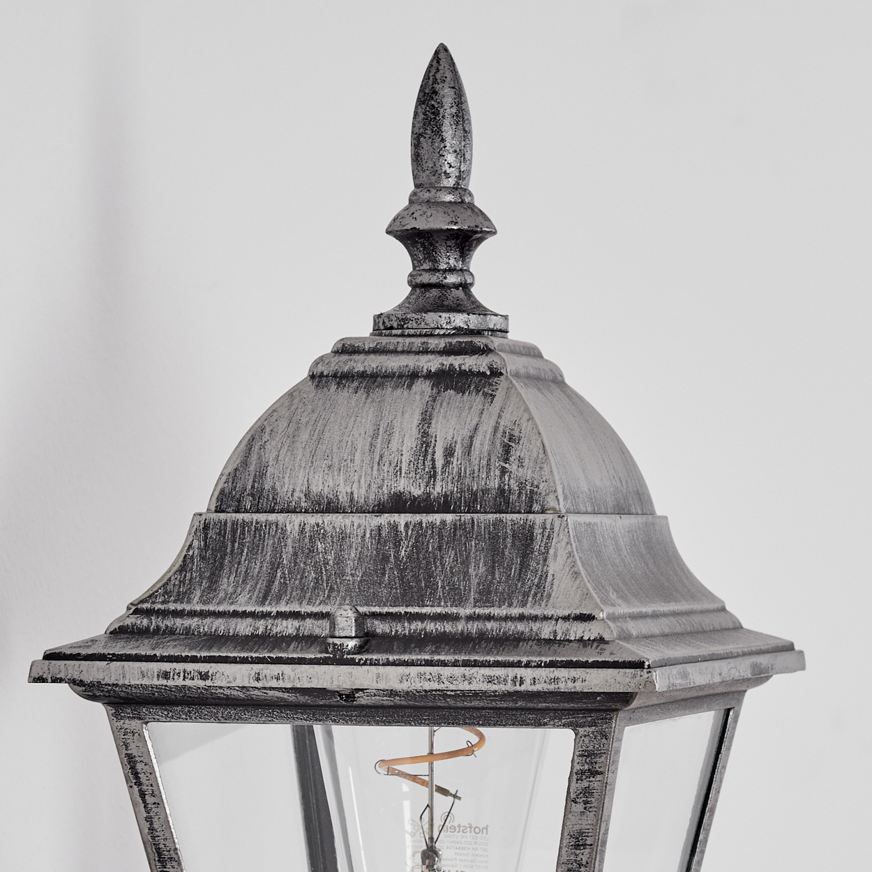 Nahaufnahme einer silbernen Außenlampe mit Glasscheibe. Die Lampe hat eine spitze Oberseite und eine Glühbirne.