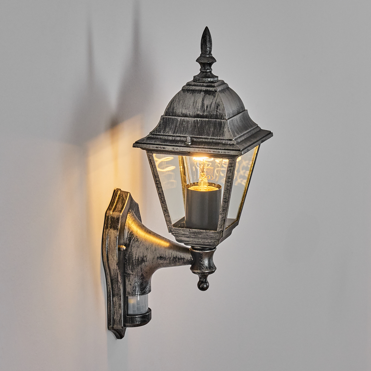 Eine Außenwandleuchte mit einer leuchtenden Glühbirne im Inneren. Die Lampe ist an einer weißen Wand befestigt.