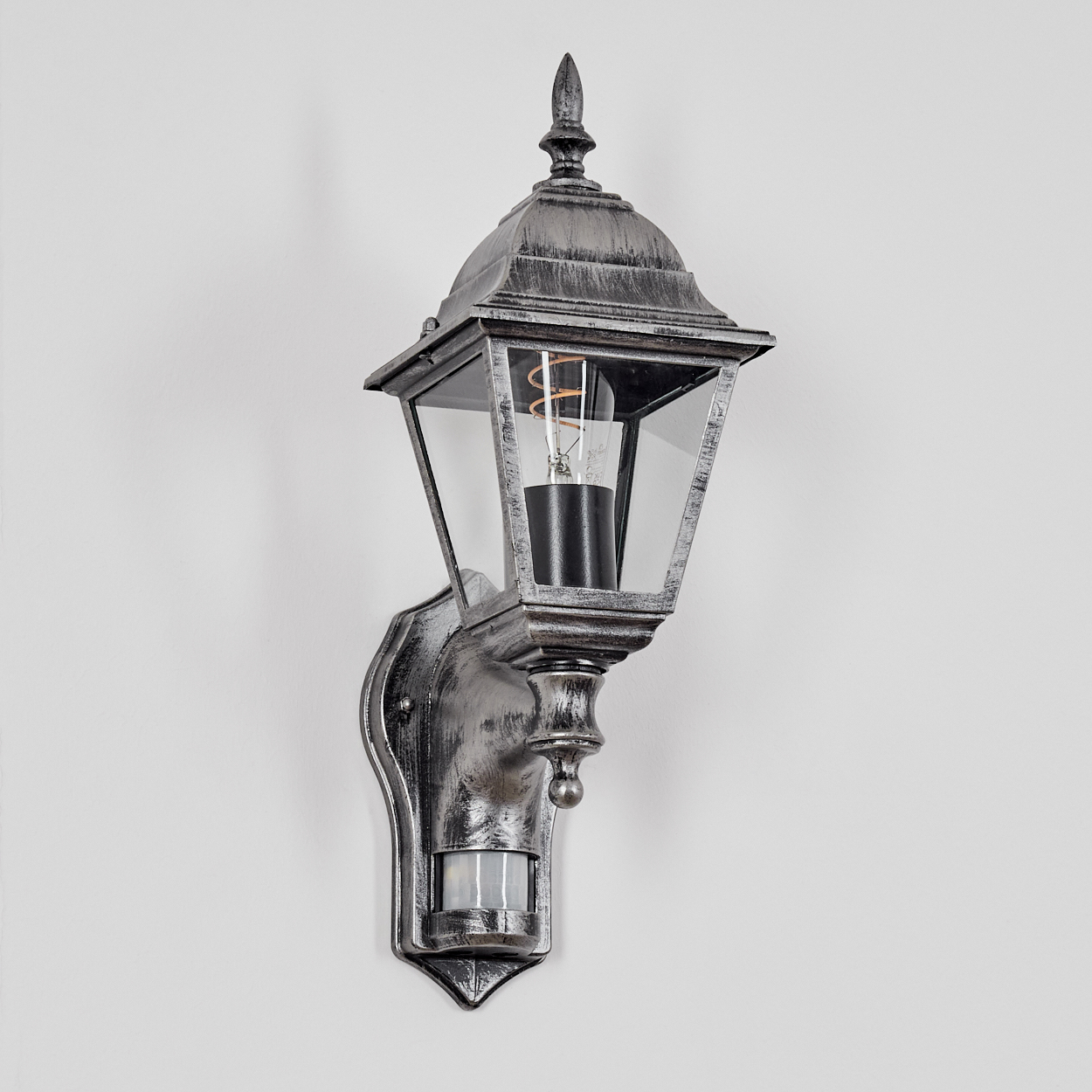 Eine metallgraue Außenlampe mit Glasgehäuse, Glühbirne und Bewegungssensor, an einer weißen Wand montiert.