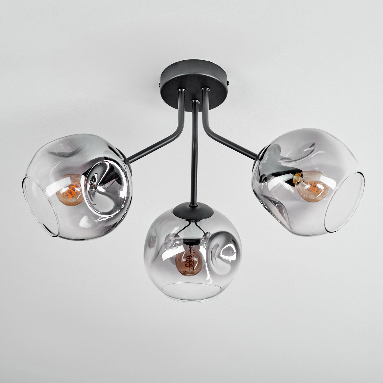Eine moderne Deckenleuchte mit drei rauchigen Glasschirmen, jede mit einer sichtbaren Glühbirne, an einem schwarzen Metallrahmen befestigt.