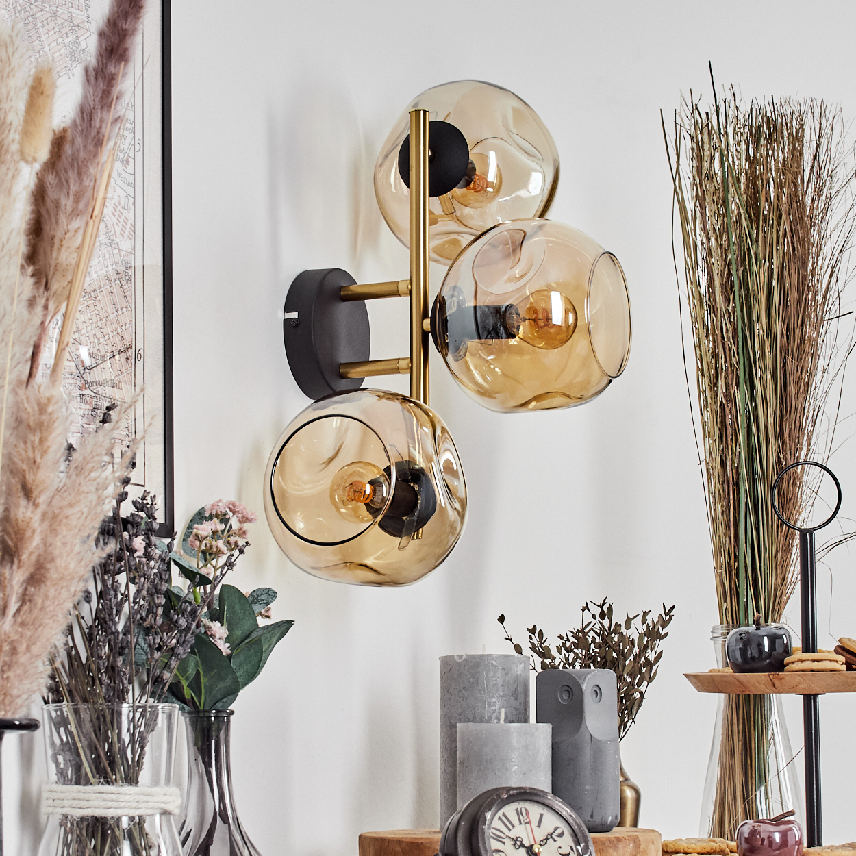 Eine Wandlampe mit drei Glaskugeln. Vor der weißen Wand befinden sich verschiedene Dekorationen, darunter Kerzen, Blumen und Uhren.