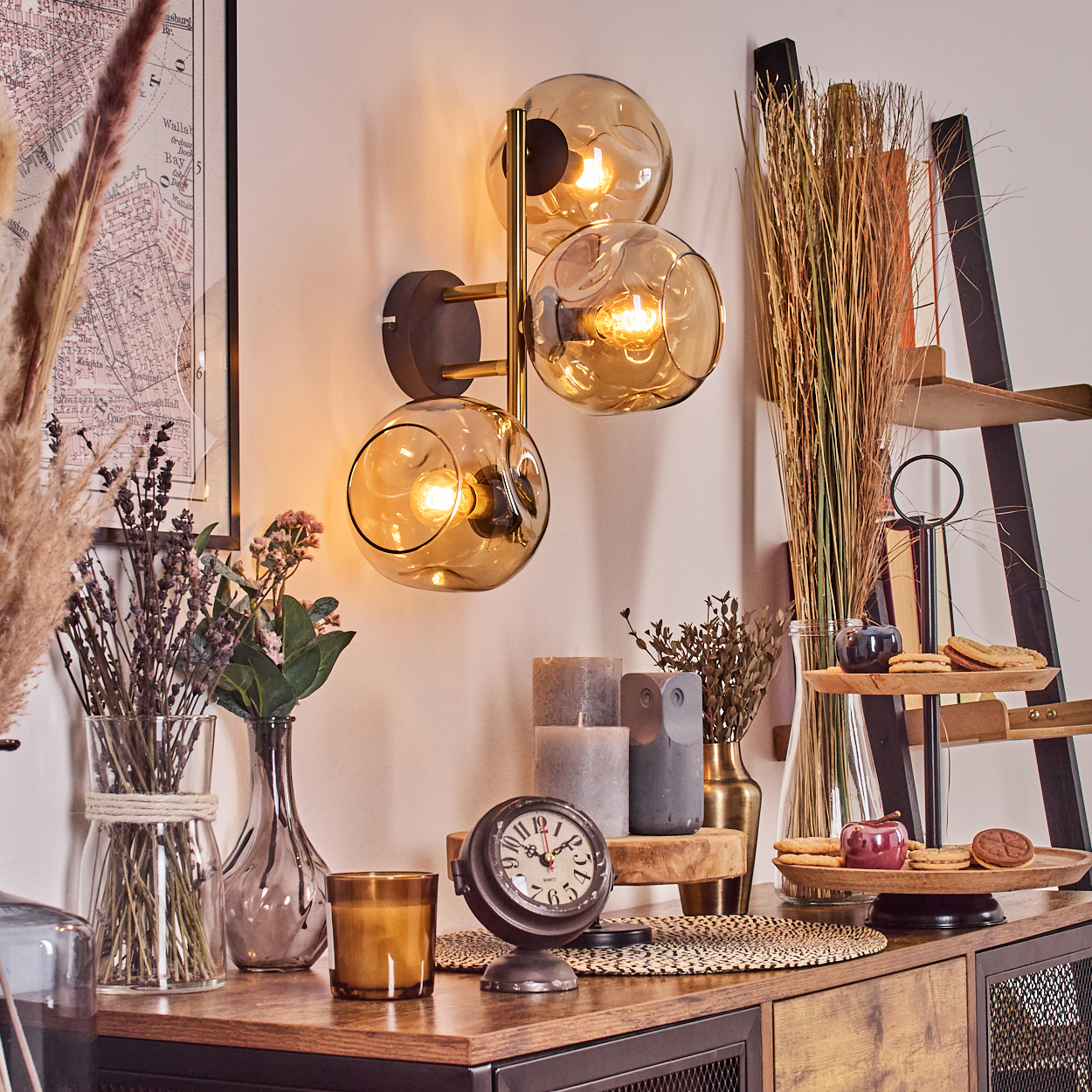 Eine dekorative Szene mit Lampe, Blumen, Kerzen, einer Uhr und einem Leiternregal mit Keksen und Früchten.