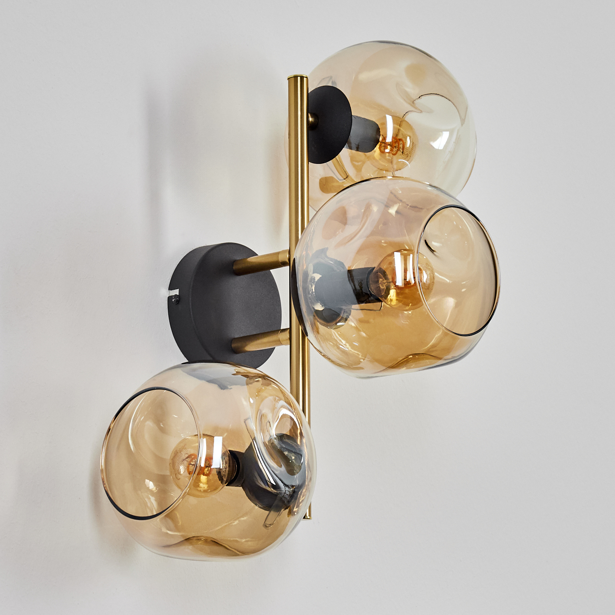 Eine moderne Wandleuchte mit drei kugelförmigen, bernsteinfarbenen Glasschirmen. Sie hat schwarze und goldfarbene Metallkomponenten. Der Hintergrund ist eine schlichte weiße Wand.