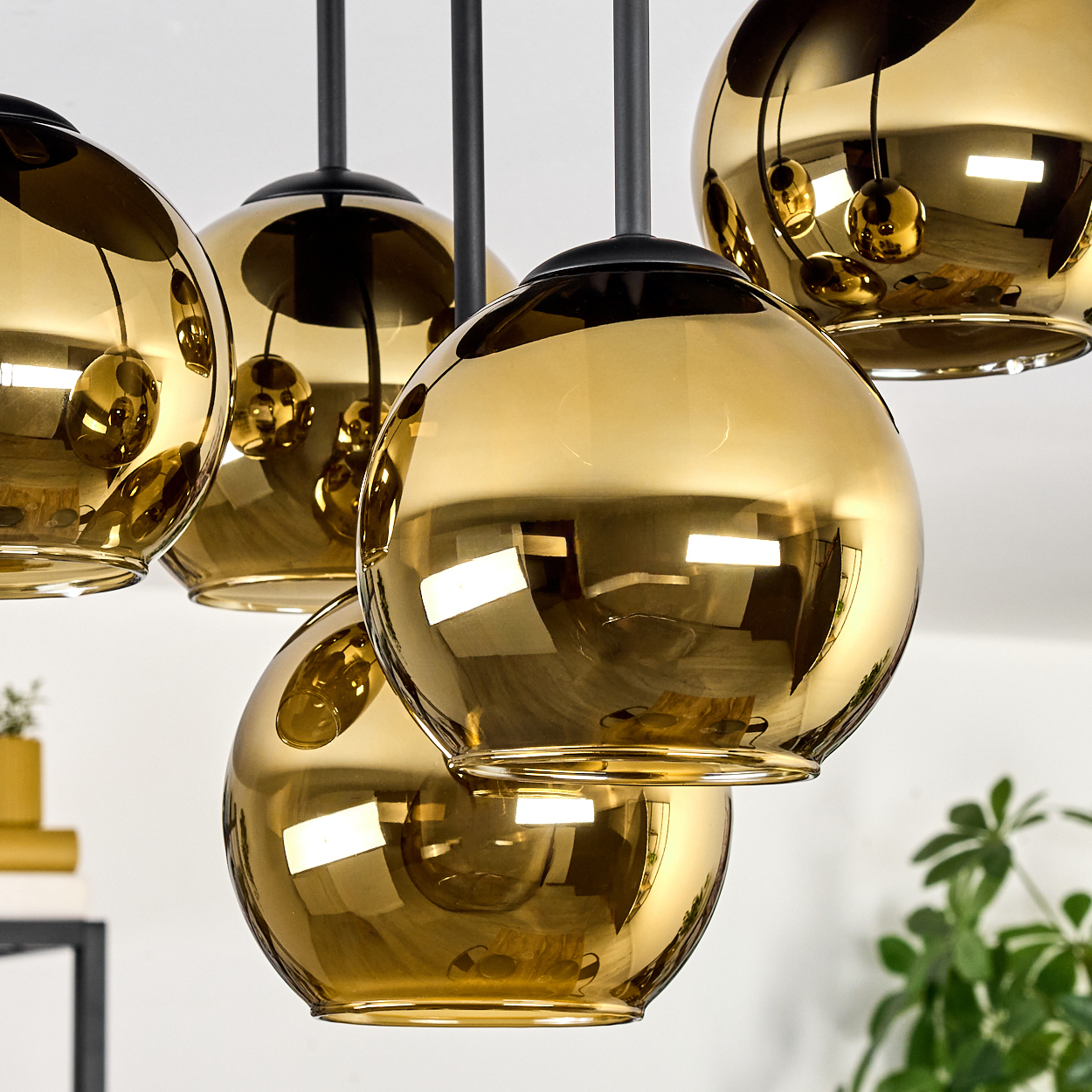 Nahaufnahme eines modernen Kronleuchters mit mehreren goldfarbenen, kugelförmigen Glasschirmen, die an schwarzen Stäben hängen. Im Hintergrund ist eine Pflanze.