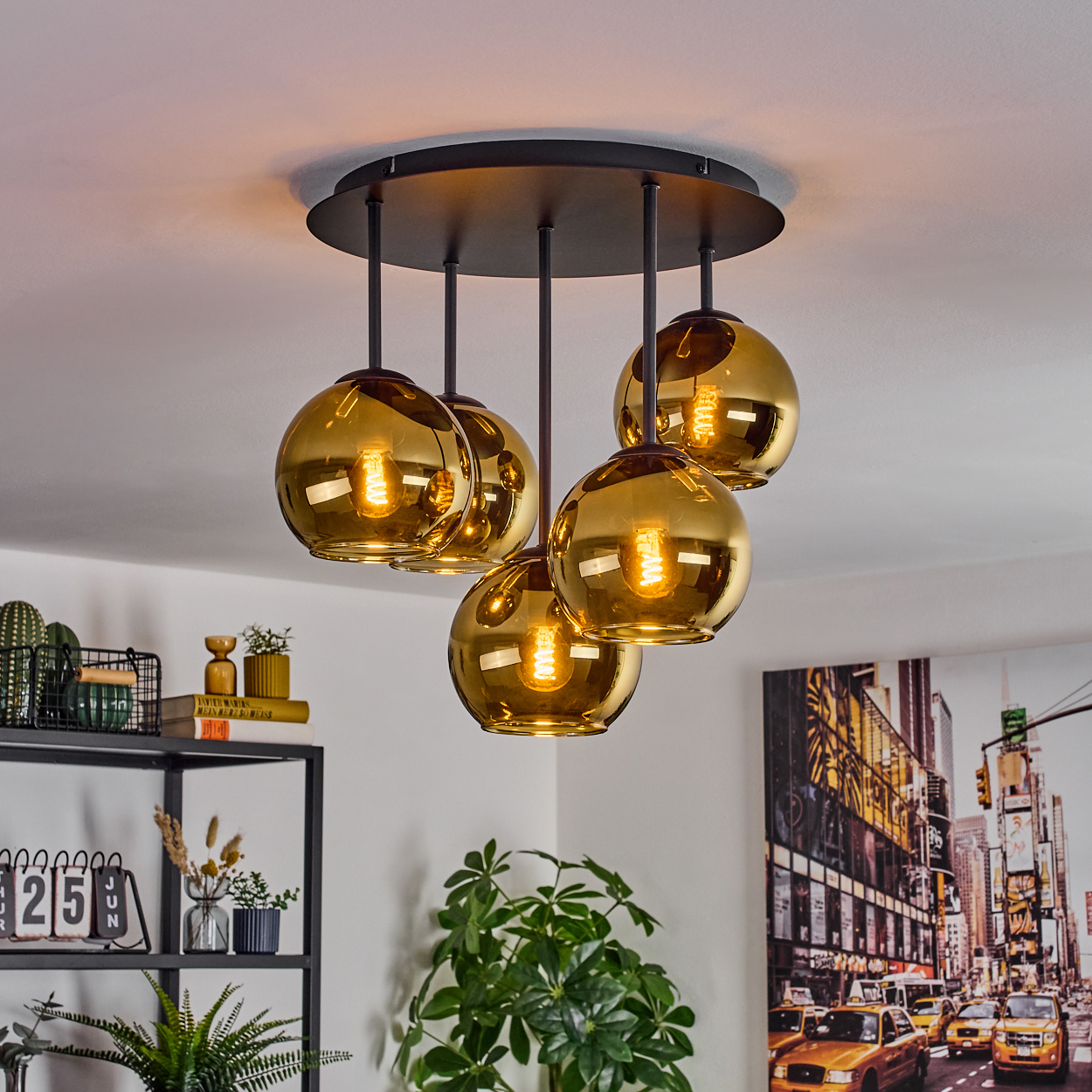 Eine moderne Deckenleuchte mit fünf goldenen Glaskugeln hängt an einer schwarzen Basis. Ein Stadtbildgemälde und Pflanzen sind sichtbar.