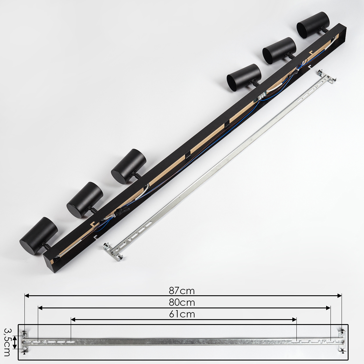 Eine schwarze Leuchte mit sechs zylindrischen Strahlern. Sie liegt auf einer weißen Oberfläche, zusammen mit einer metallischen Halterung.