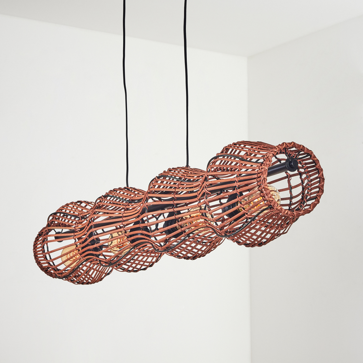Eine braune Pendelleuchte aus Korbgeflecht hängt an schwarzen Schnüren. Sie hat ein wellenförmiges Design mit sichtbaren Glühbirnen vor einer weißen Wand.