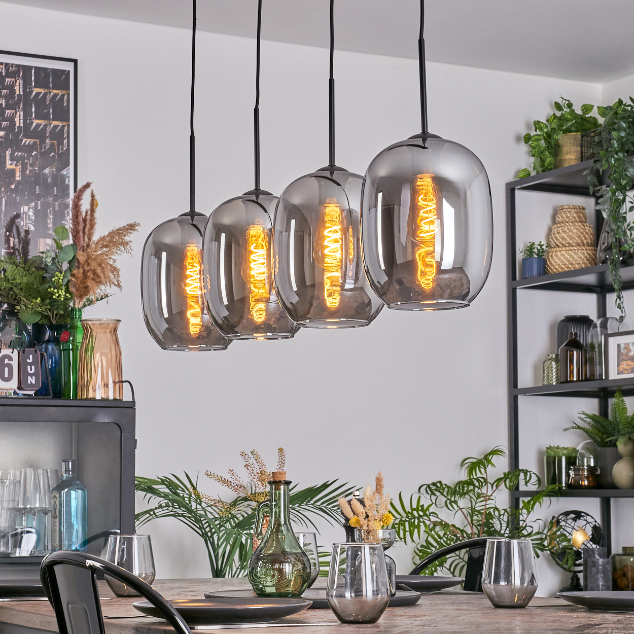 Esszimmer mit vier Pendelleuchten. Tisch mit Gläsern und Tellern. Regale mit Pflanzen und Dekoration im Hintergrund.
