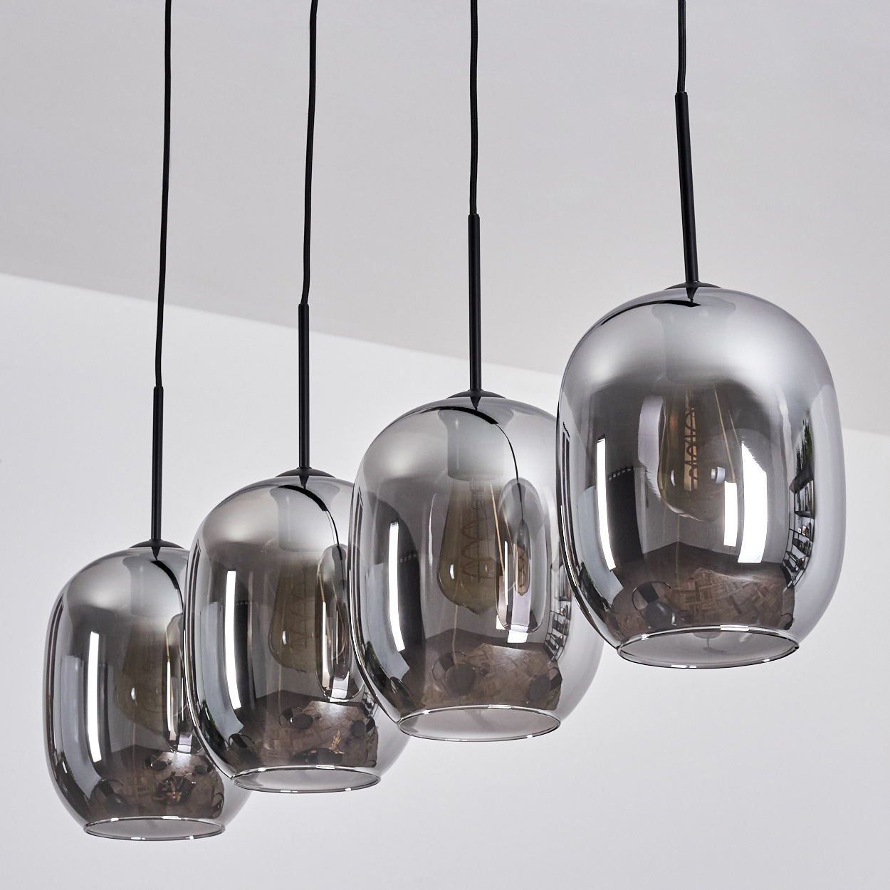 Vier graue Glas-Pendelleuchten hängen an schwarzen Kabeln von einer weißen Decke. Jede Glühbirne ist im Glas sichtbar.