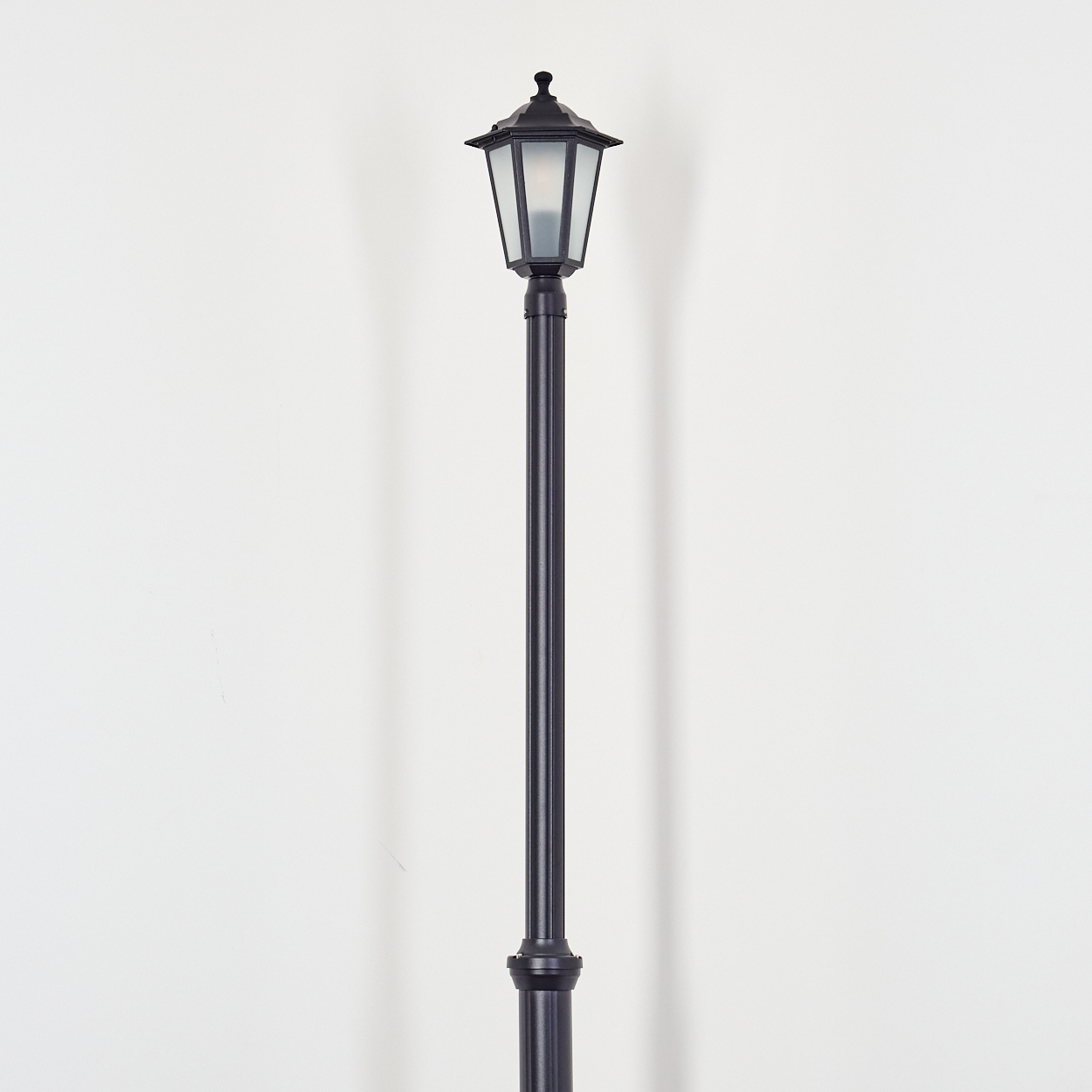 Eine schwarze Straßenlaterne steht vor einer weißen Wand. Die Lampe hat einen Laternenaufsatz und eine hohe Stange.