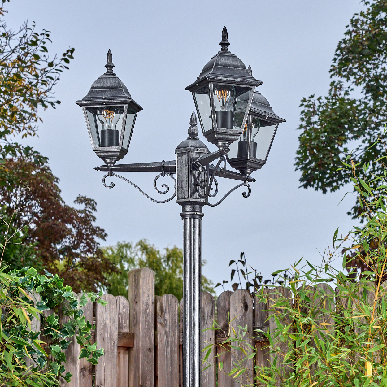 Eine dekorative Laterne mit drei Lampen steht in einem Garten. Im Hintergrund sind ein Holzzaun und grüne Pflanzen zu sehen.
