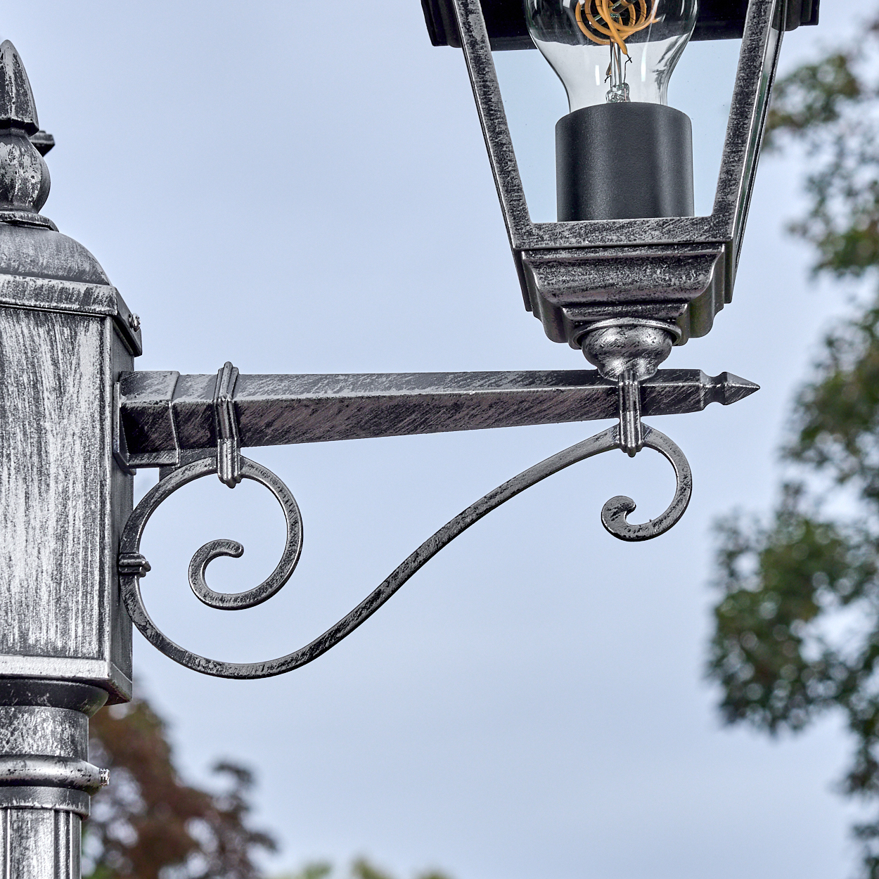 Nahaufnahme einer dekorativen Laterne mit Metallrahmen. Die Lampe hat eine Glühbirne, der Hintergrund ist bewölkt und Bäume.