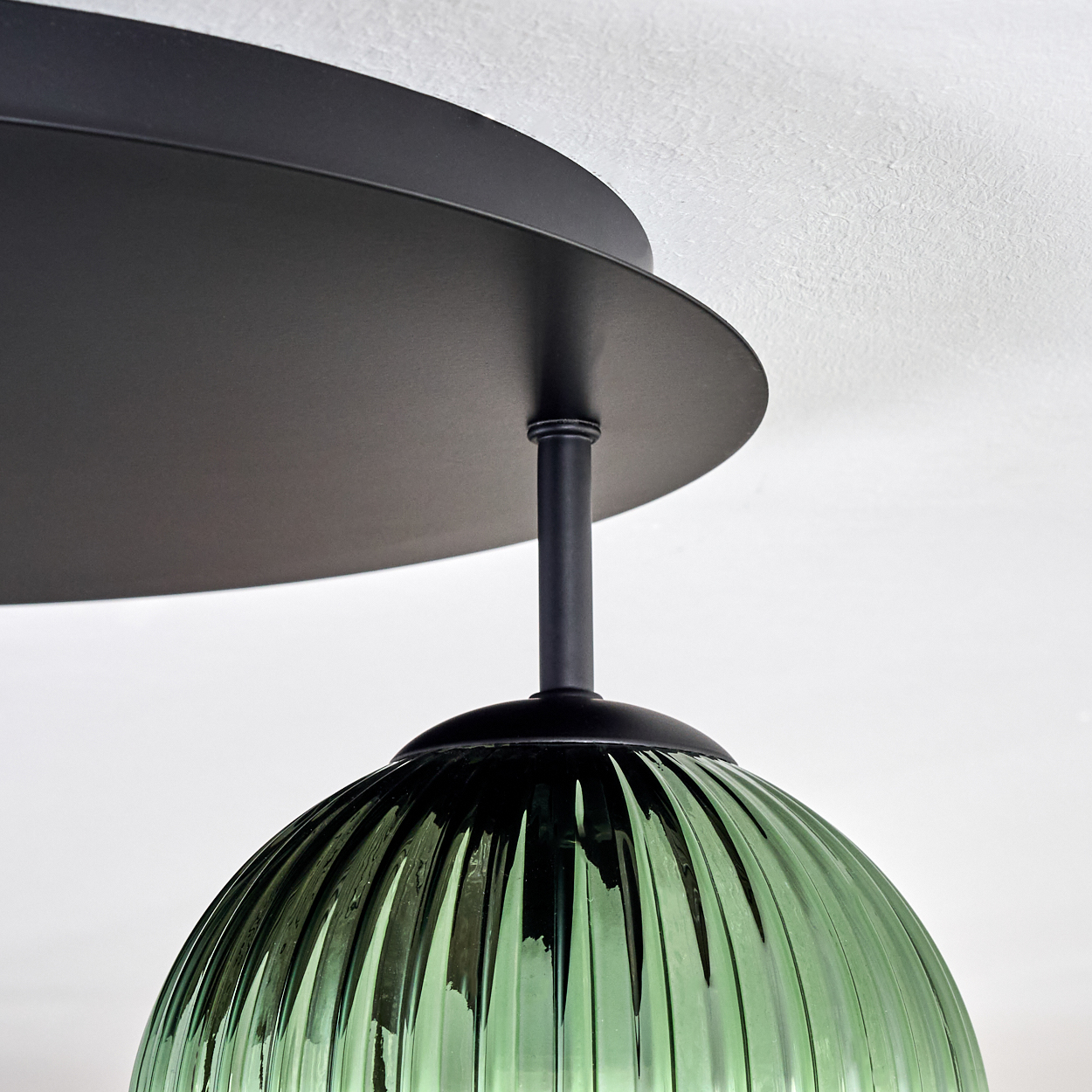 Eine moderne Deckenlampe mit schwarzem, kreisförmigem Sockel und grünem, geripptem Glasschirm. Der Hintergrund ist eine weiße Decke.