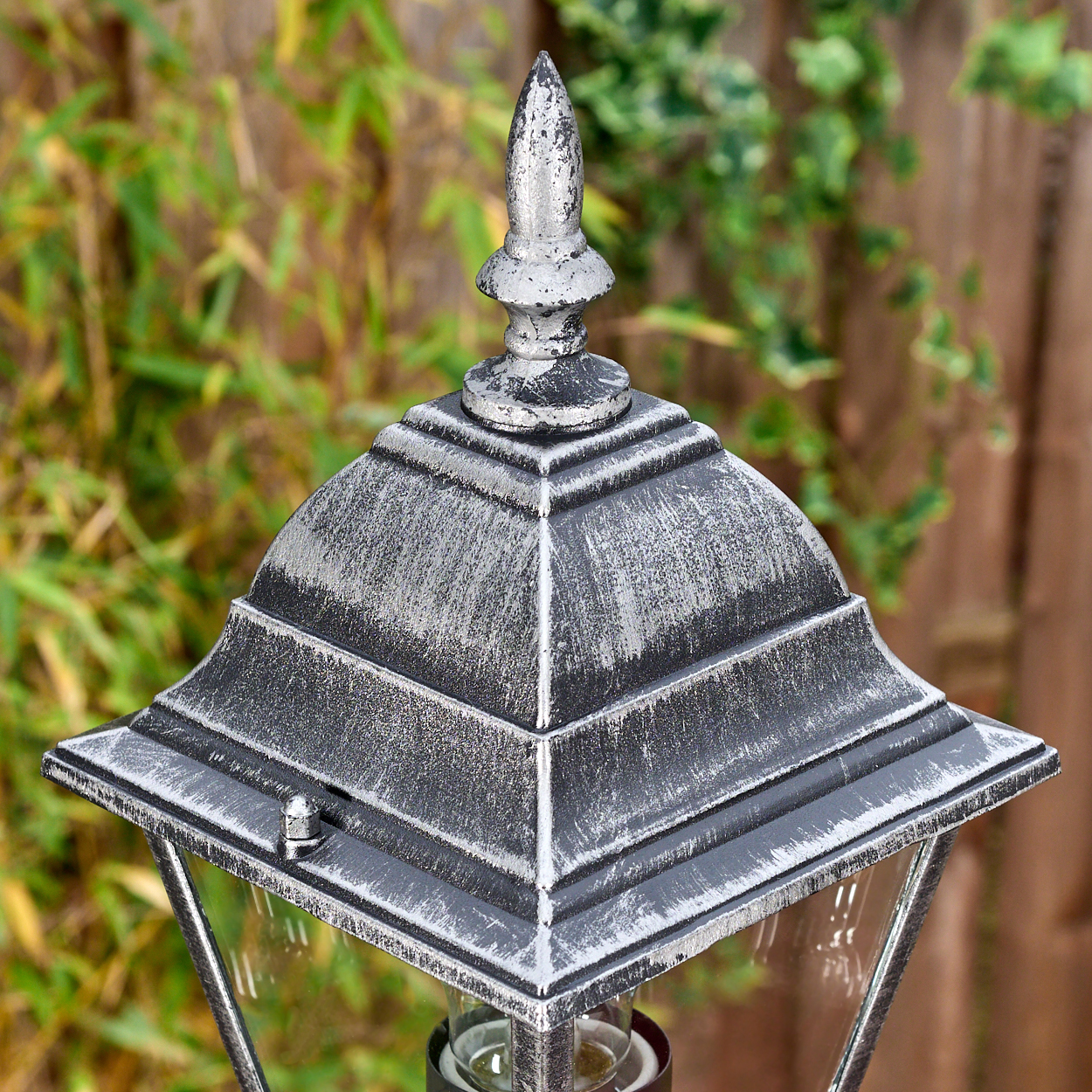 Nahaufnahme einer silbernen Außenlampe. Die Lampe hat eine spitze Oberseite und einen quadratischen Körper mit Glasscheiben. Grünes Laub und ein Holzzaun sind im Hintergrund.