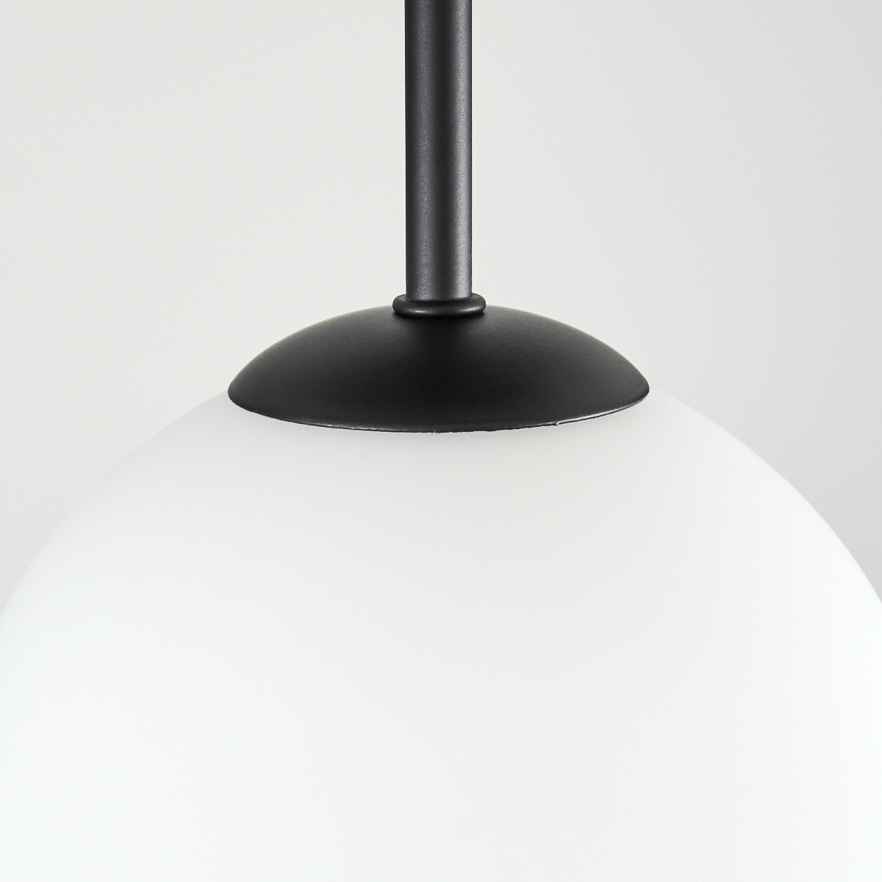 Nahaufnahme einer schwarzen Pendelleuchte. Sie hat einen weißen, runden Glasschirm. Die Lampe befindet sich vor weißem Hintergrund.