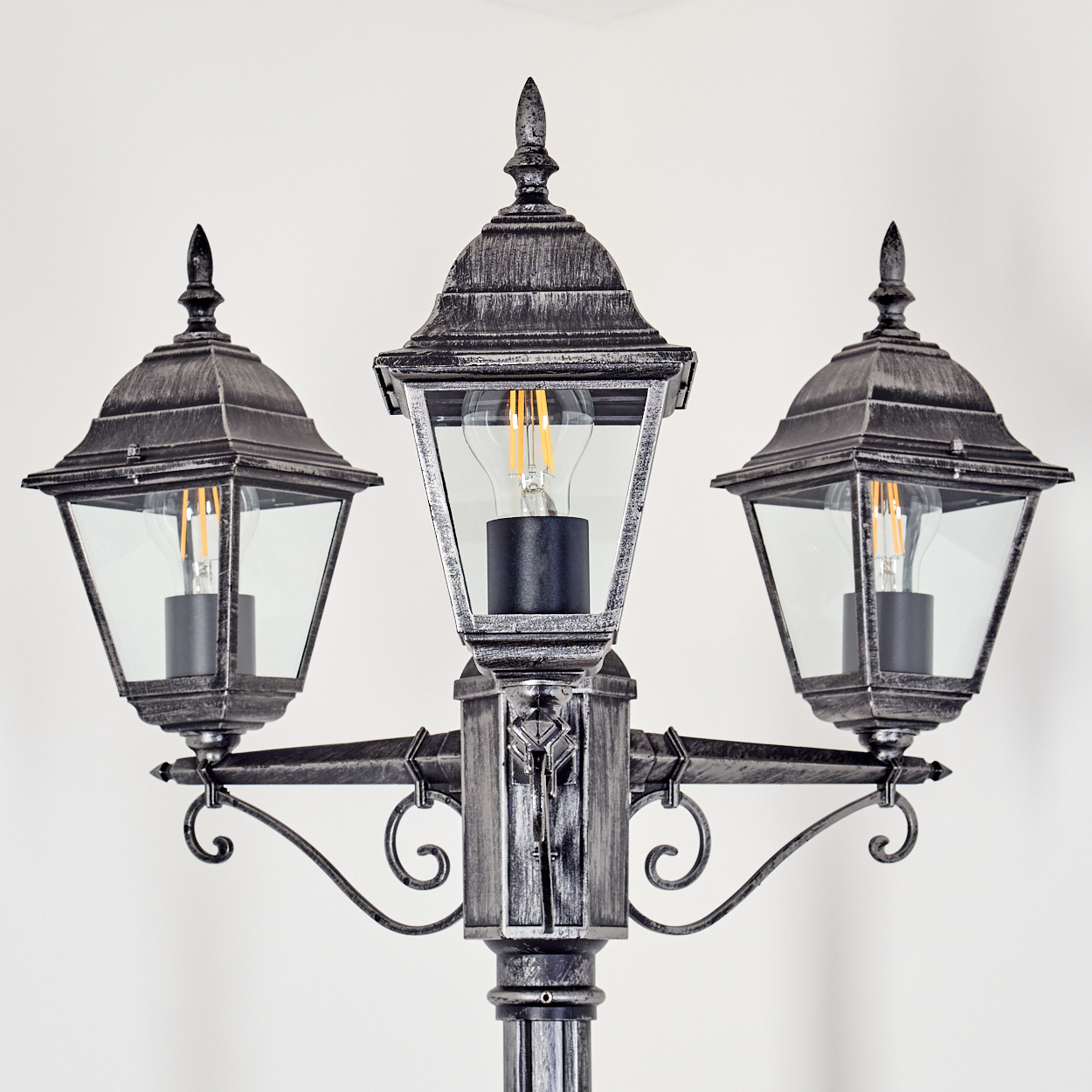 Nahaufnahme einer dreilampigen Straßenlaterne. Die Lampen sind verziert, grau und halten jeweils eine leuchtende Glühbirne vor weißem Hintergrund.