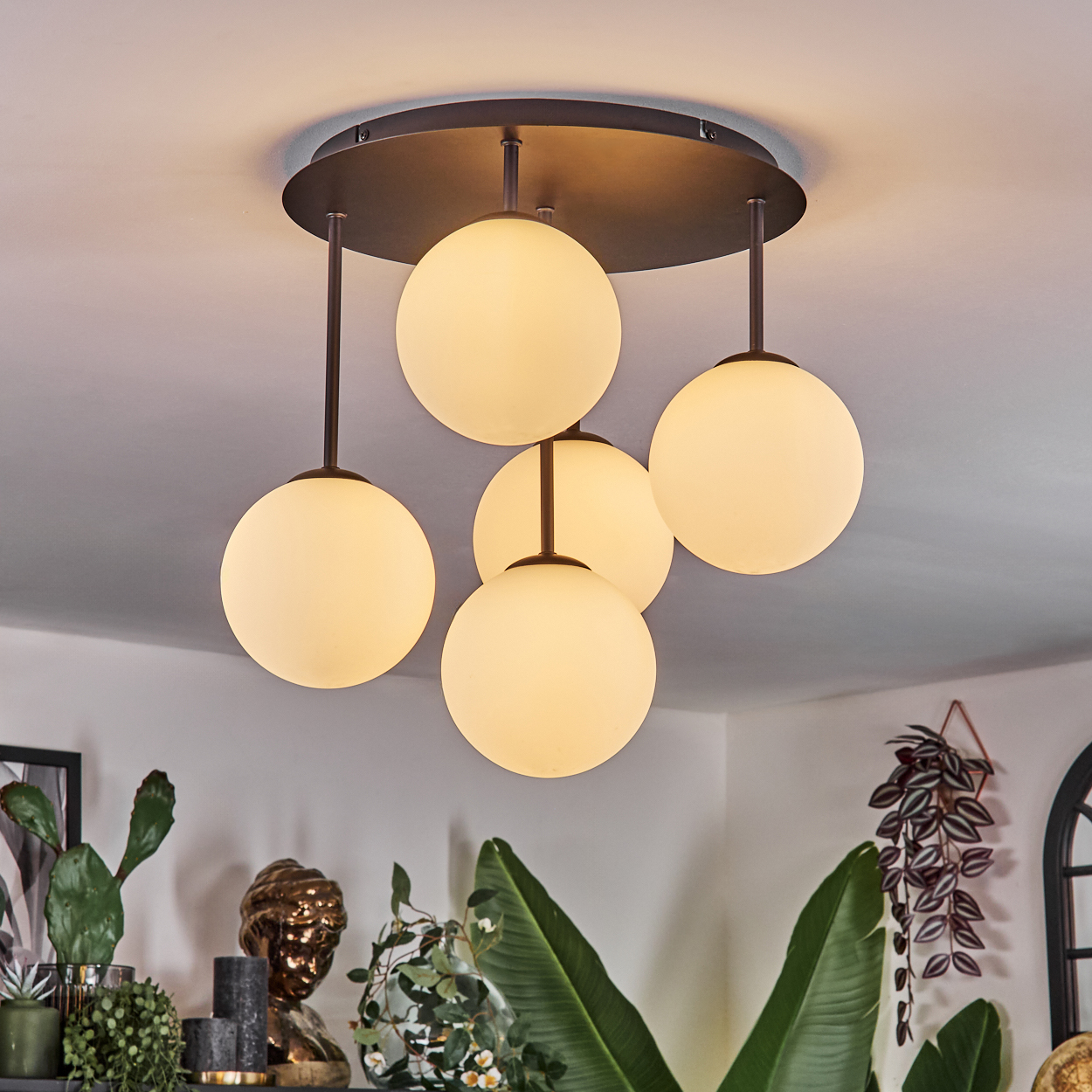 Ein moderner Kronleuchter mit fünf kugelförmigen Lichtern hängt von der Decke. Dekorative Pflanzen und eine Büste sind im Hintergrund zu sehen.