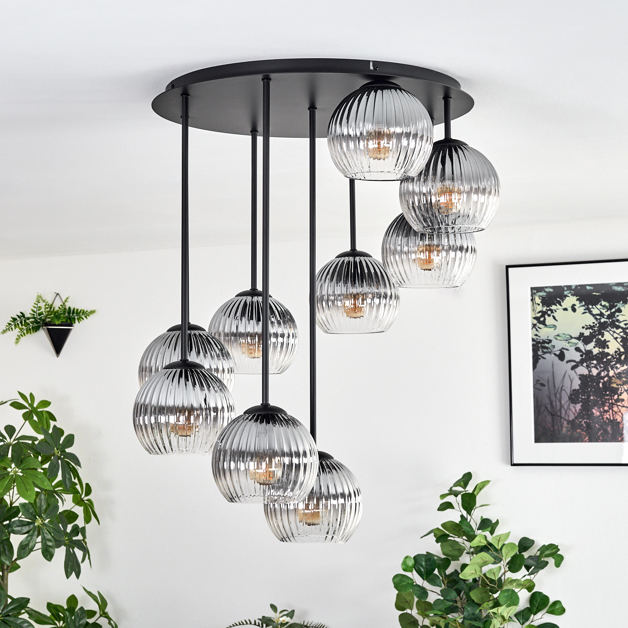 Eine moderne Deckenlampe mit mehreren kugelförmigen Lichtern hängt von einer weißen Decke. Grüne Pflanzen und ein gerahmtes Bild schmücken die Wand.