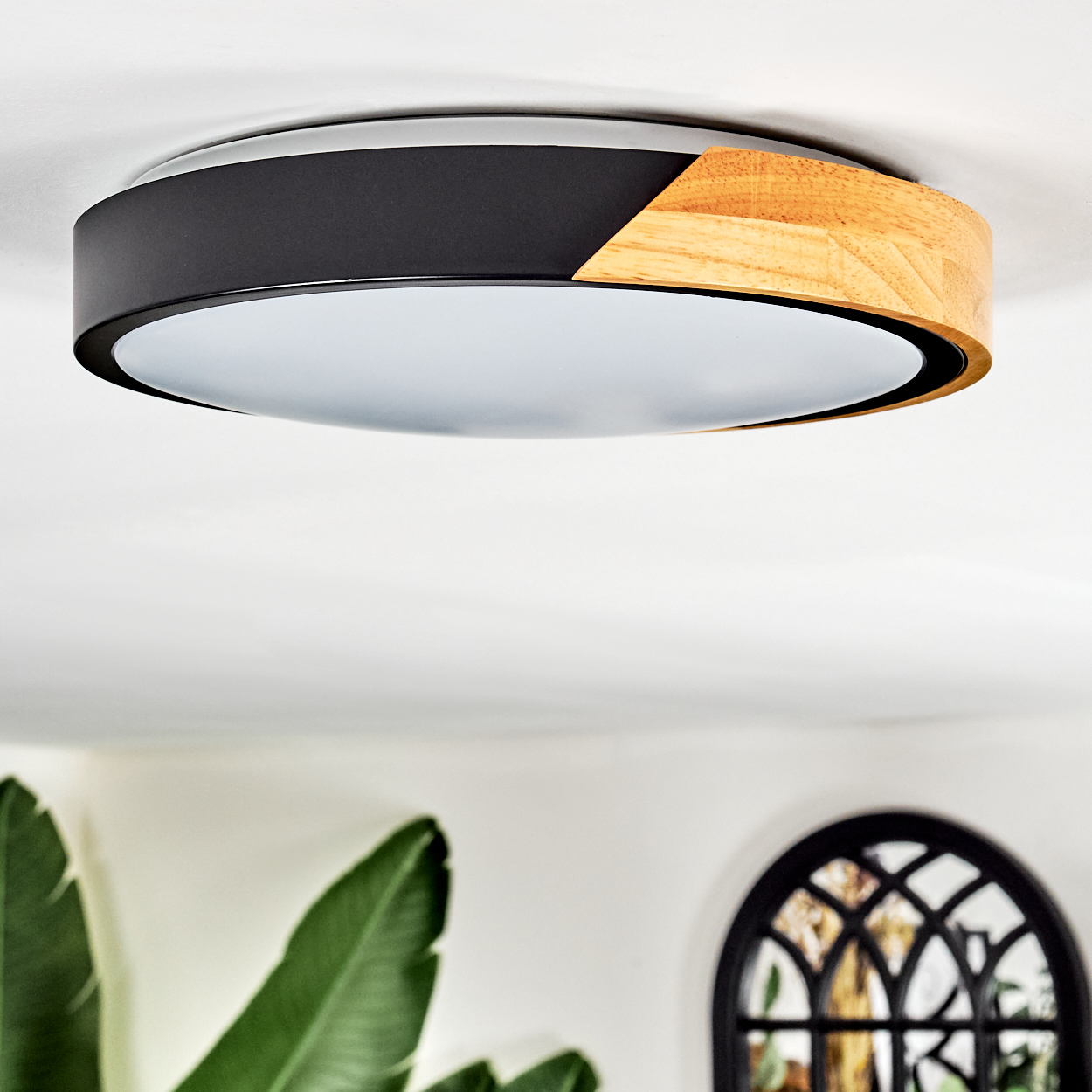 Eine moderne Deckenlampe mit schwarzem und Holzrahmen. Der Hintergrund zeigt eine weiße Wand, Pflanzen und ein Fenster.