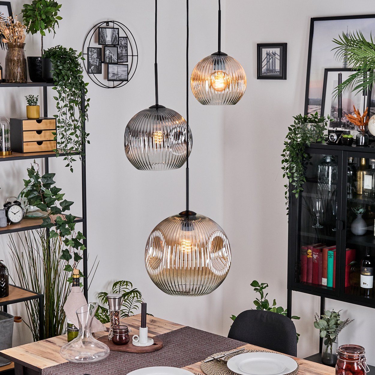 Esszimmer mit drei Hängelampen. Tisch mit Tellern gedeckt. Regale mit Pflanzen, Uhr und Dekorationen. Schwarz gerahmte Bilder.