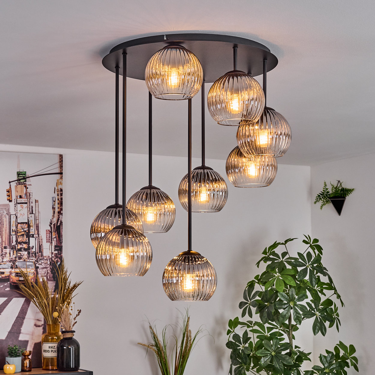 Eine moderne Deckenleuchte mit neun Glaskugelleuchten hängt von der Decke. Im Vordergrund steht eine Pflanze, an der Wand hängt ein Stadtbild.