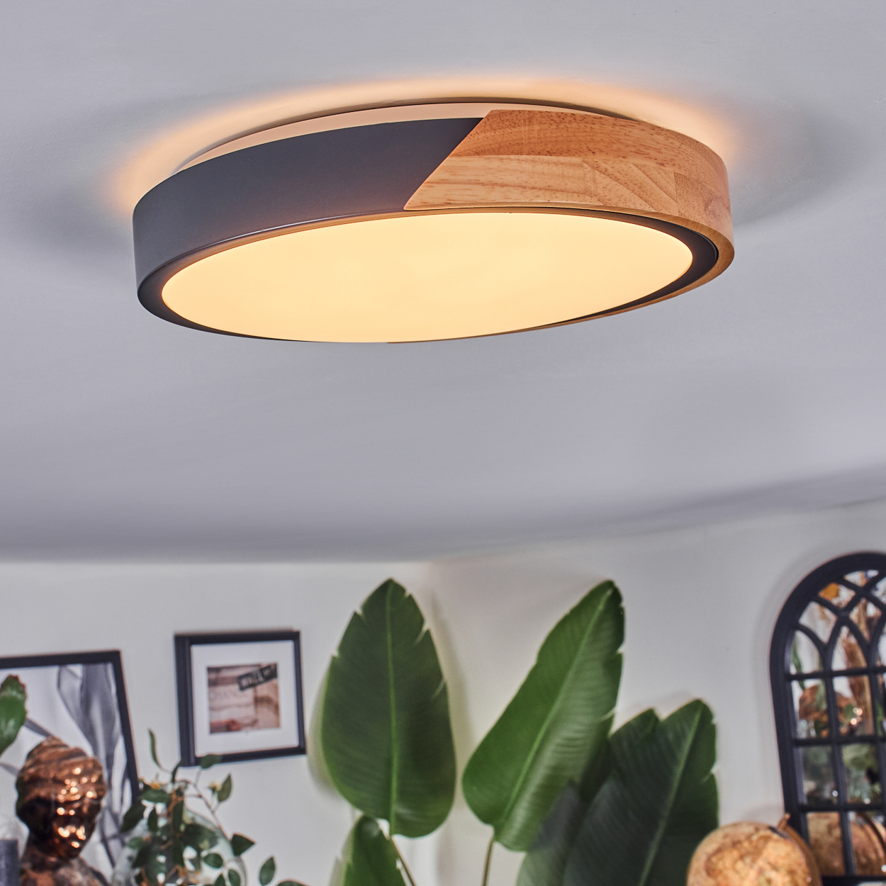 Eine moderne Deckenleuchte mit Holz- und dunkelgrauen Akzenten. Im Hintergrund grüne Pflanzen und Bilder. Das Licht ist an.
