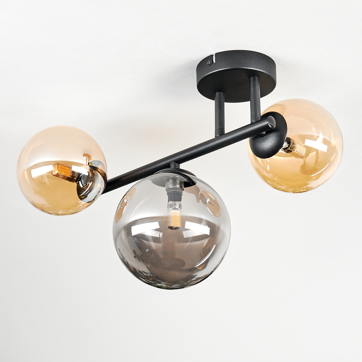 Moderne Deckenleuchte mit drei Glaskugeln. Zwei bernsteinfarben, eine grau. Schwarze Metallarme und Deckenhalterung. Weißer Hintergrund.
