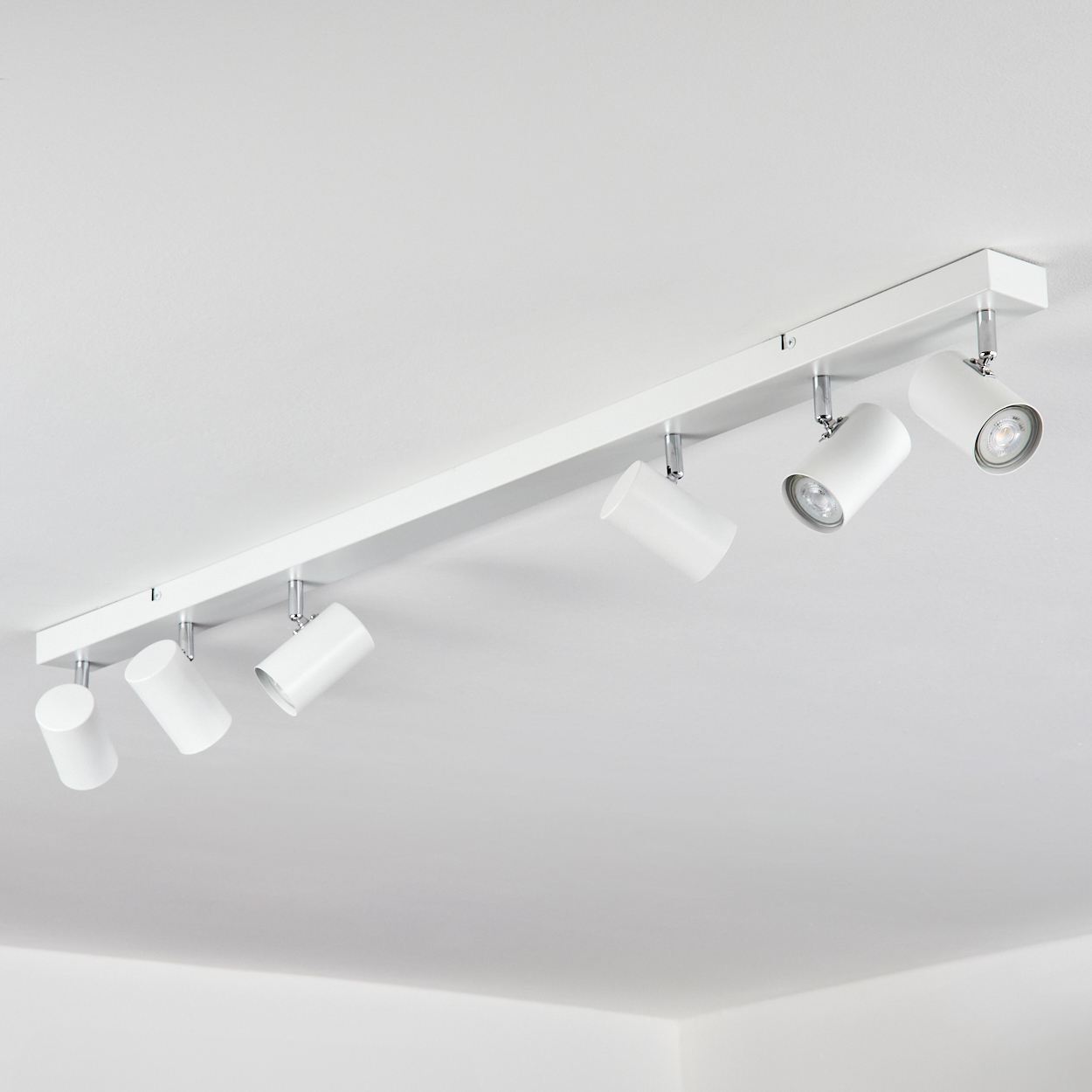 Weißer Deckenstrahler mit sechs verstellbaren Lichtern. Er ist auf einer weißen rechteckigen Basis montiert. Die Lichter sind nach unten gerichtet.