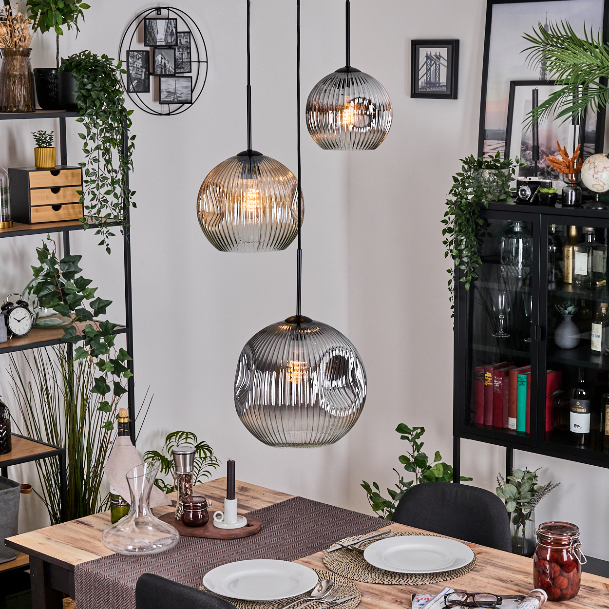 Esszimmer mit Holztisch, drei Hängelampen, Tellern und einem schwarzen Schrank mit verschiedenen Objekten und Pflanzen.