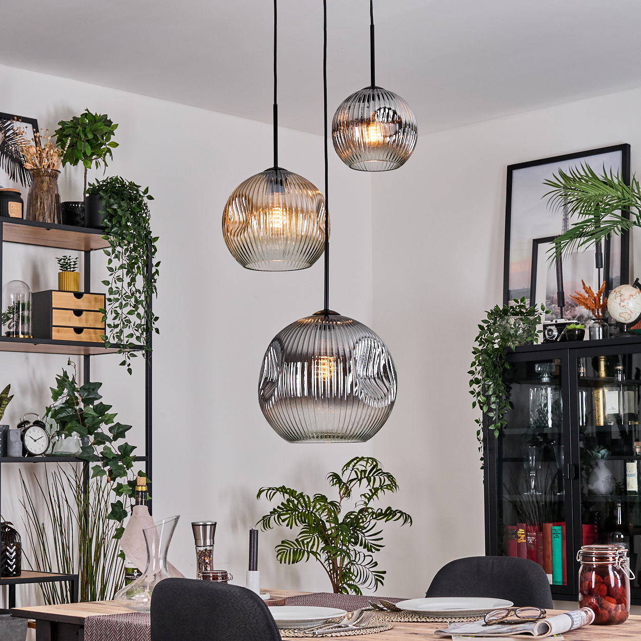 Drei Glaskugel-Pendelleuchten hängen über einem Esstisch, der für eine Mahlzeit gedeckt ist. Regale und ein Schrank sind im Hintergrund.